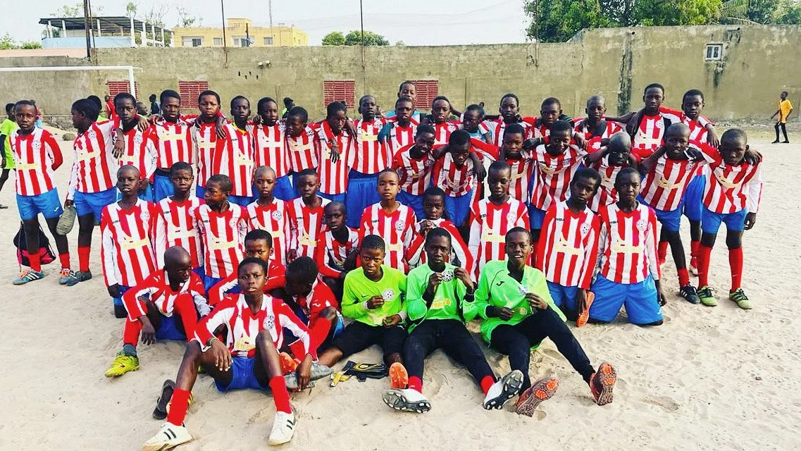 A group of young boys in soccer uniforms, red and white striped shirts and blue shorts, posing on a sandy field. Some are standing, some kneeling, and a few sitting in the front. Four boys in green goalkeeper jerseys are sitting in front holding soccer balls. A concrete wall and some buildings are visible in the background.