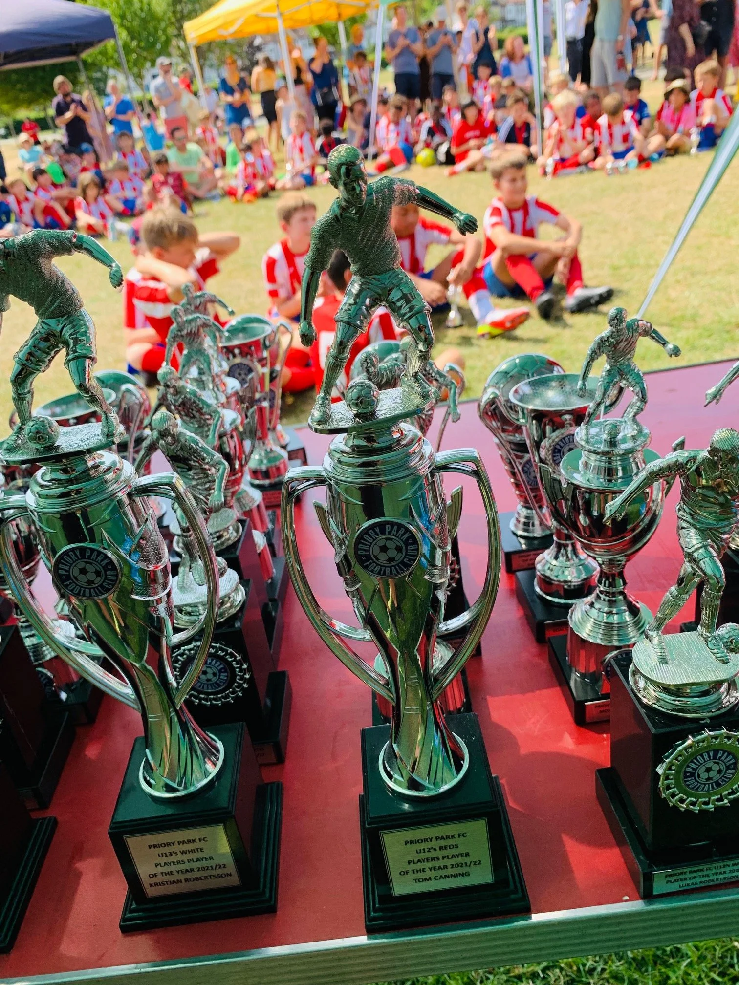 Close-up of multiple shiny silver trophies with soccer player figures on top, displayed on a red table at an outdoor event, with children and adults sitting and standing in the background.