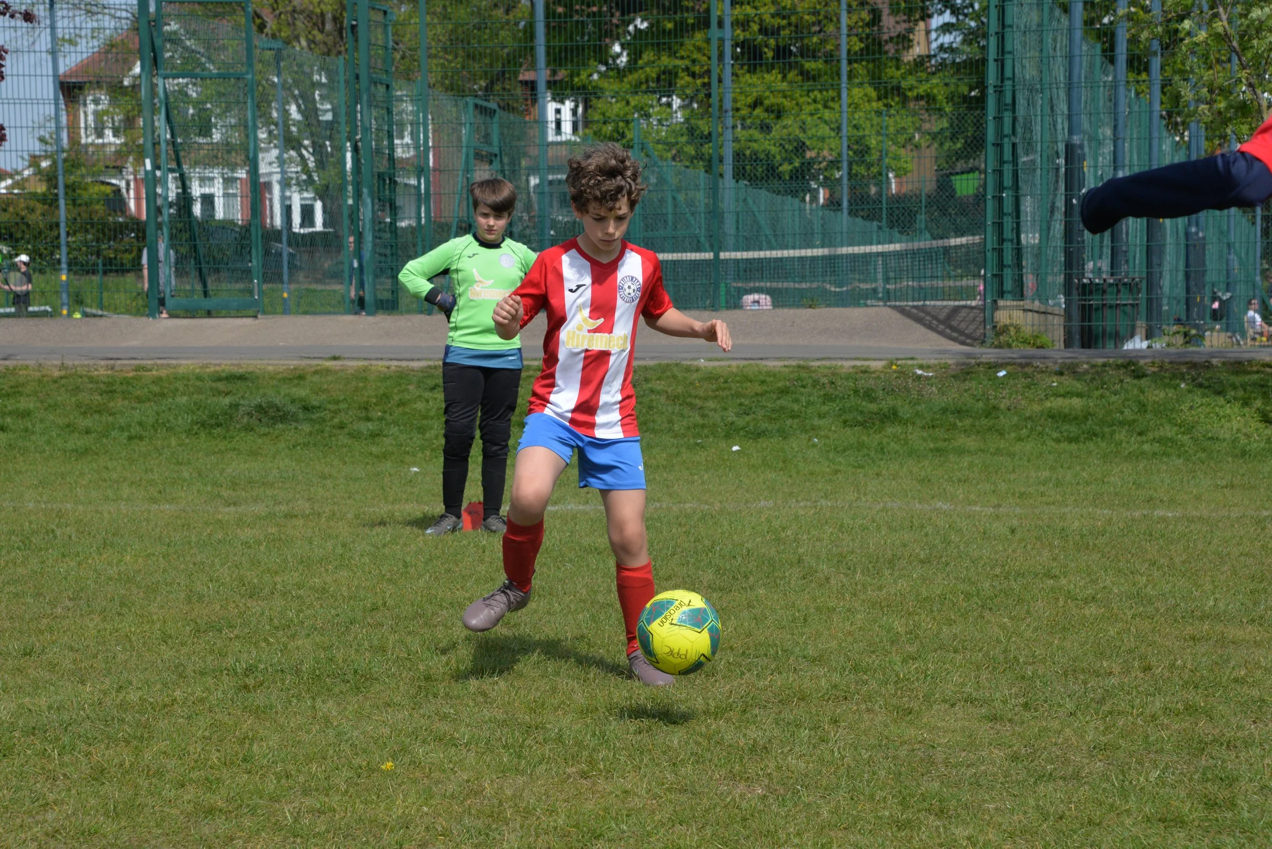 Young boy in a red and white striped soccer jersey and blue shorts kicks a yellow and green soccer ball on a grass field, with a goalkeeper and a fence in the background.