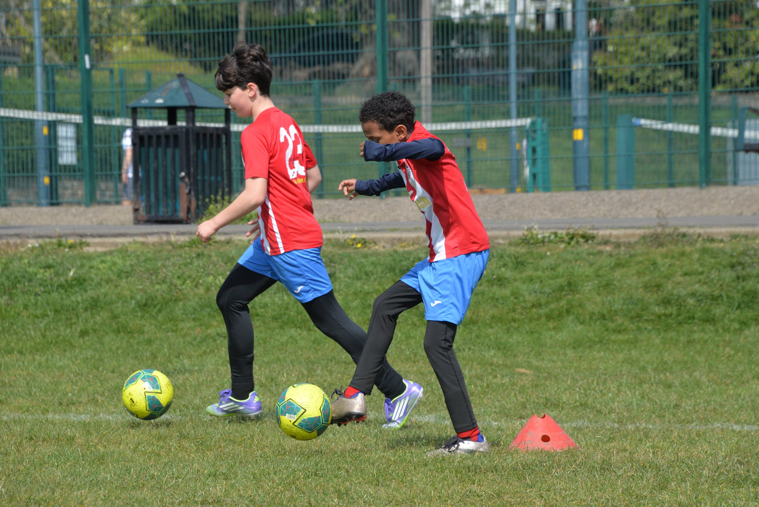 Two young boys practicing soccer on a grassy field, dribbling soccer balls and passing through orange cones, with a green fence and trees in the background.