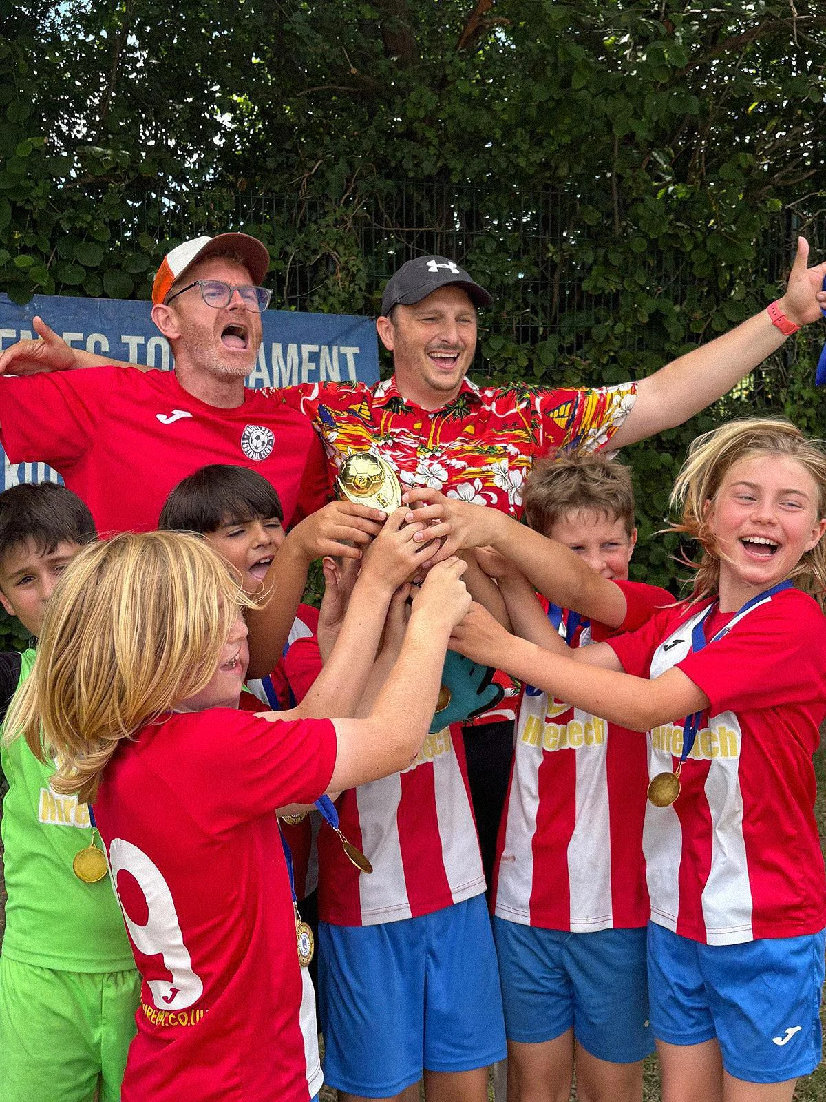 A group of children wearing red and white soccer uniforms celebrating with two coaches or adults, one holding a small trophy, as they smile and raise their arms in victory.