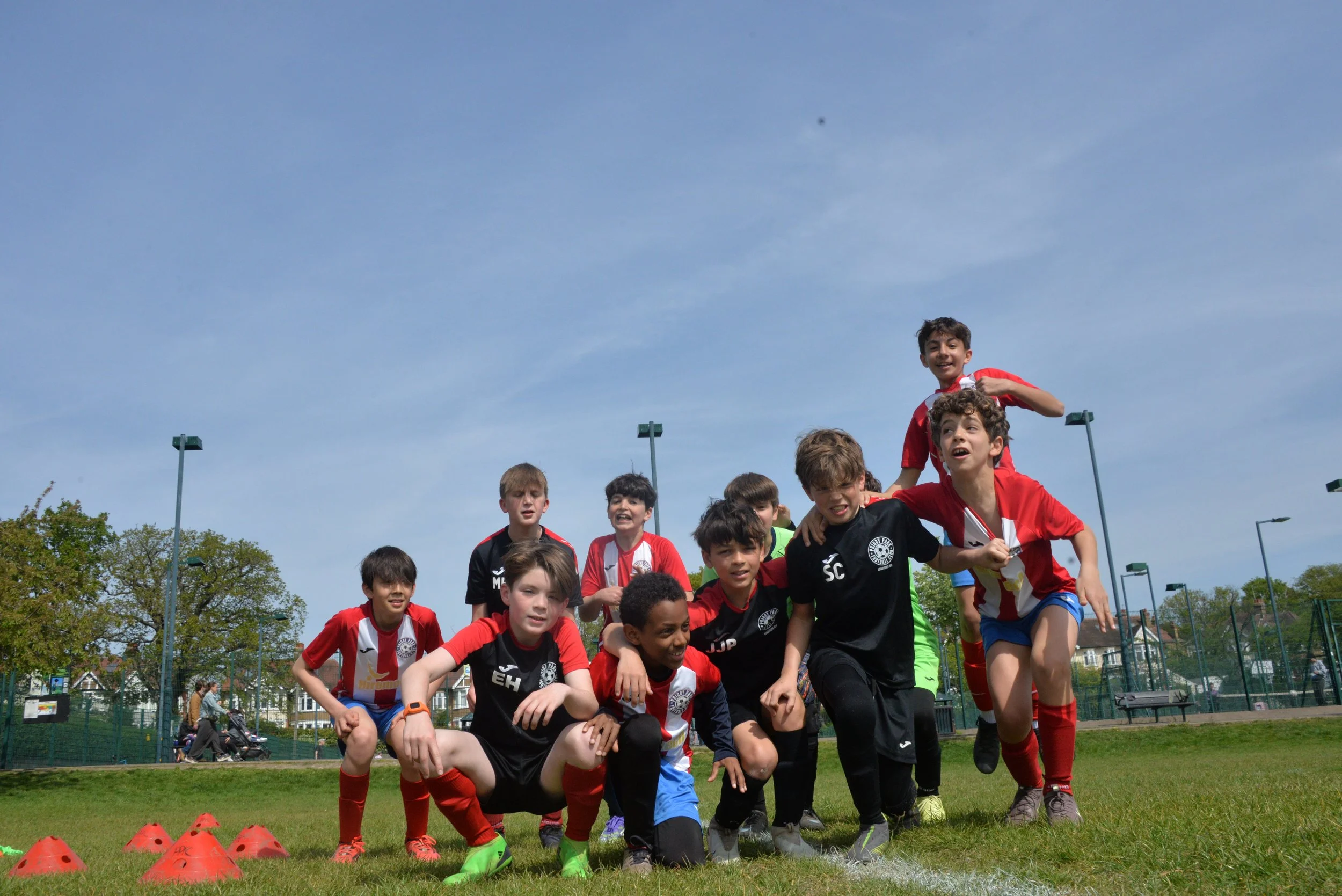 Group of young boys in sports uniforms on a soccer field, some kneeling and others standing, smiling and posing for a photo, with a playground and trees in the background under a blue sky.