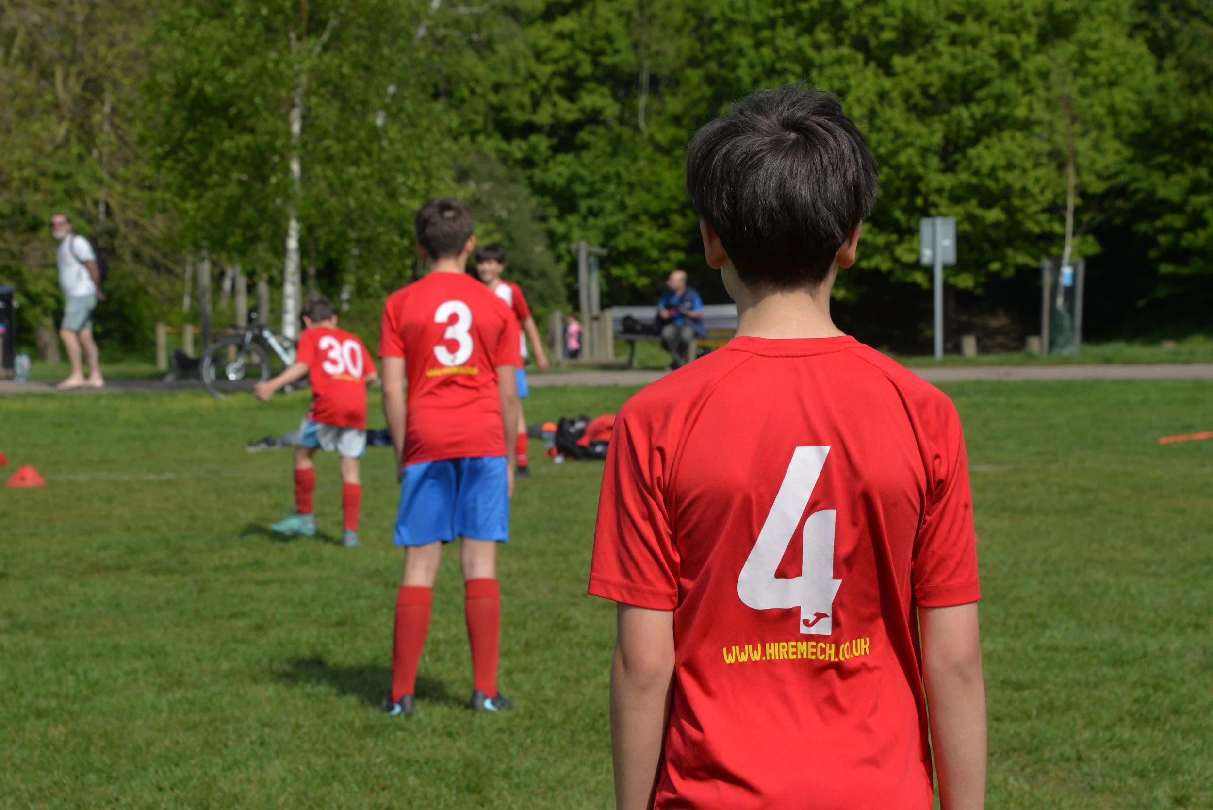 Children in red sports uniforms lined up outdoors on a grassy field, practicing or playing soccer, with green trees in the background.
