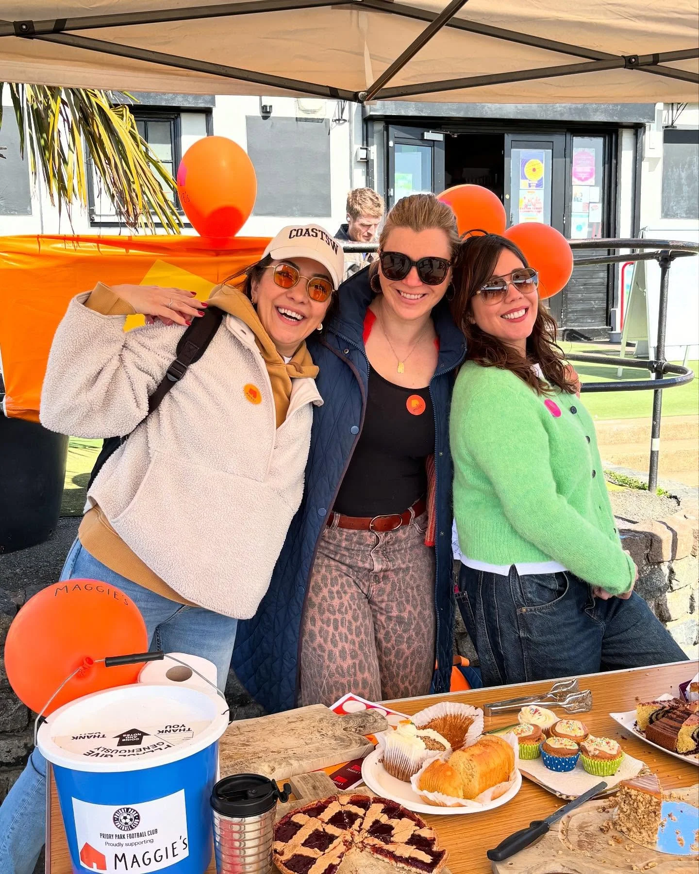 Three women smiling and posing at an outdoor gathering, with a table of desserts and orange balloons, under a canopy.