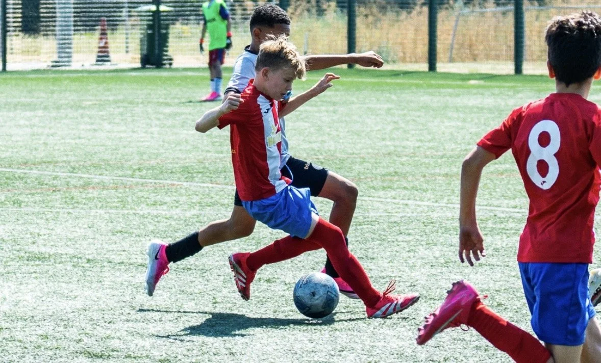 Kids playing soccer on a field, with one child in red about to kick the ball, and others around him.
