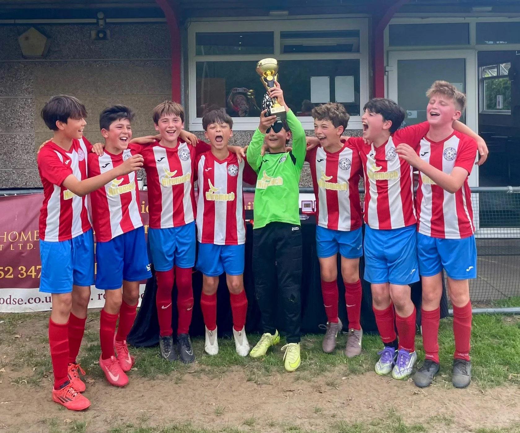 A group of young boys in red and white soccer uniforms celebrating with a trophy after a game, standing outdoors on a grassy field.