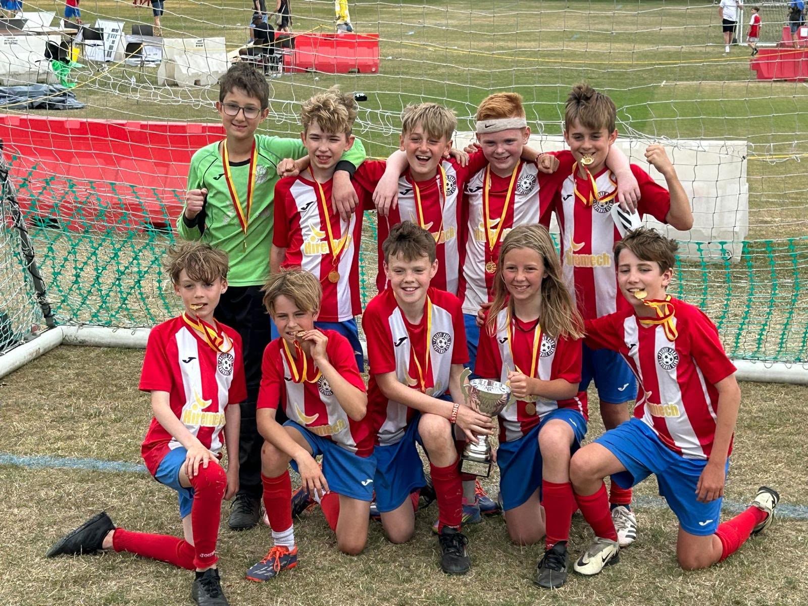 A group of young soccer players celebrating after winning a match, taking a team photo on the field. They are wearing red and white jerseys, some with medals around their necks, and are holding a trophy.