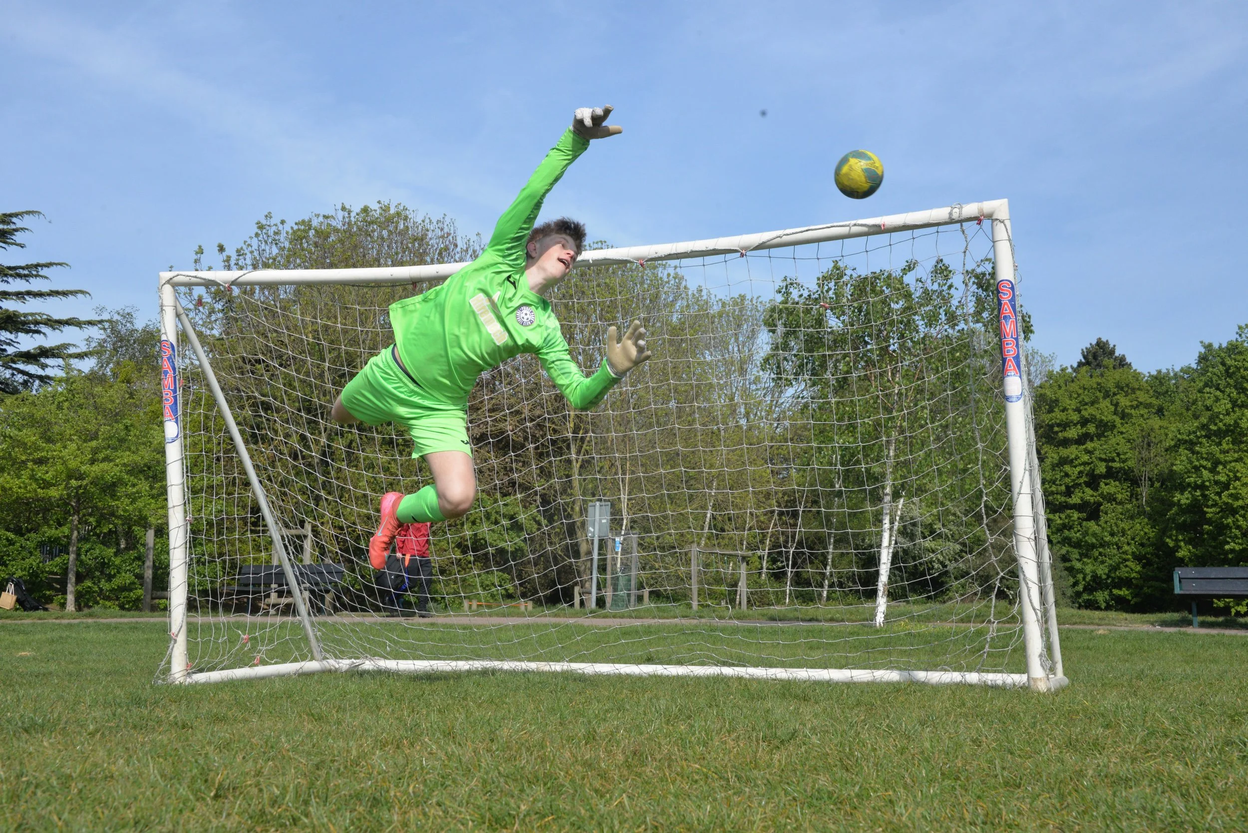 Soccer goalkeeper in green uniform diving to catch a soccer ball during a game on a field with trees in the background.