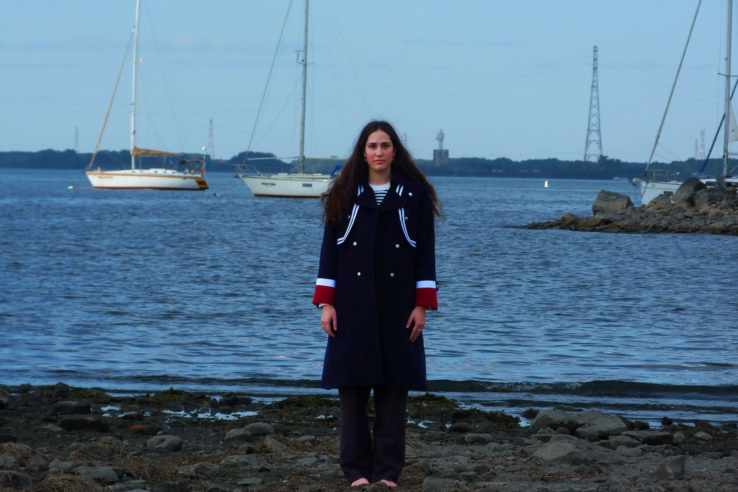 A young woman with long dark hair stands barefoot on a rocky shoreline by the water, with sailboats and a lighthouse in the background during overcast weather.