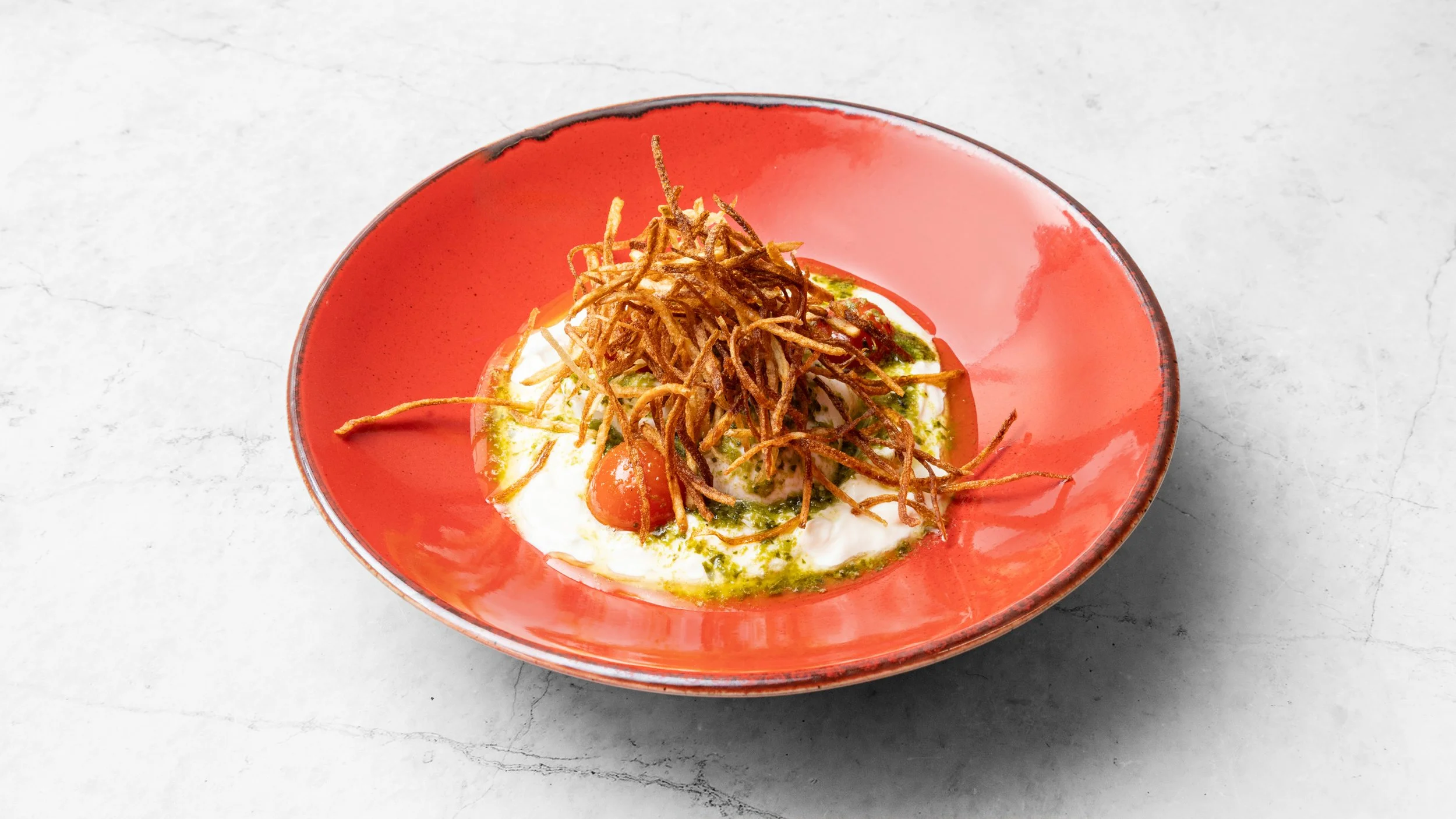 A red bowl containing soft-boiled eggs topped with crispy fried onions and green sauce, placed on a light marble surface.