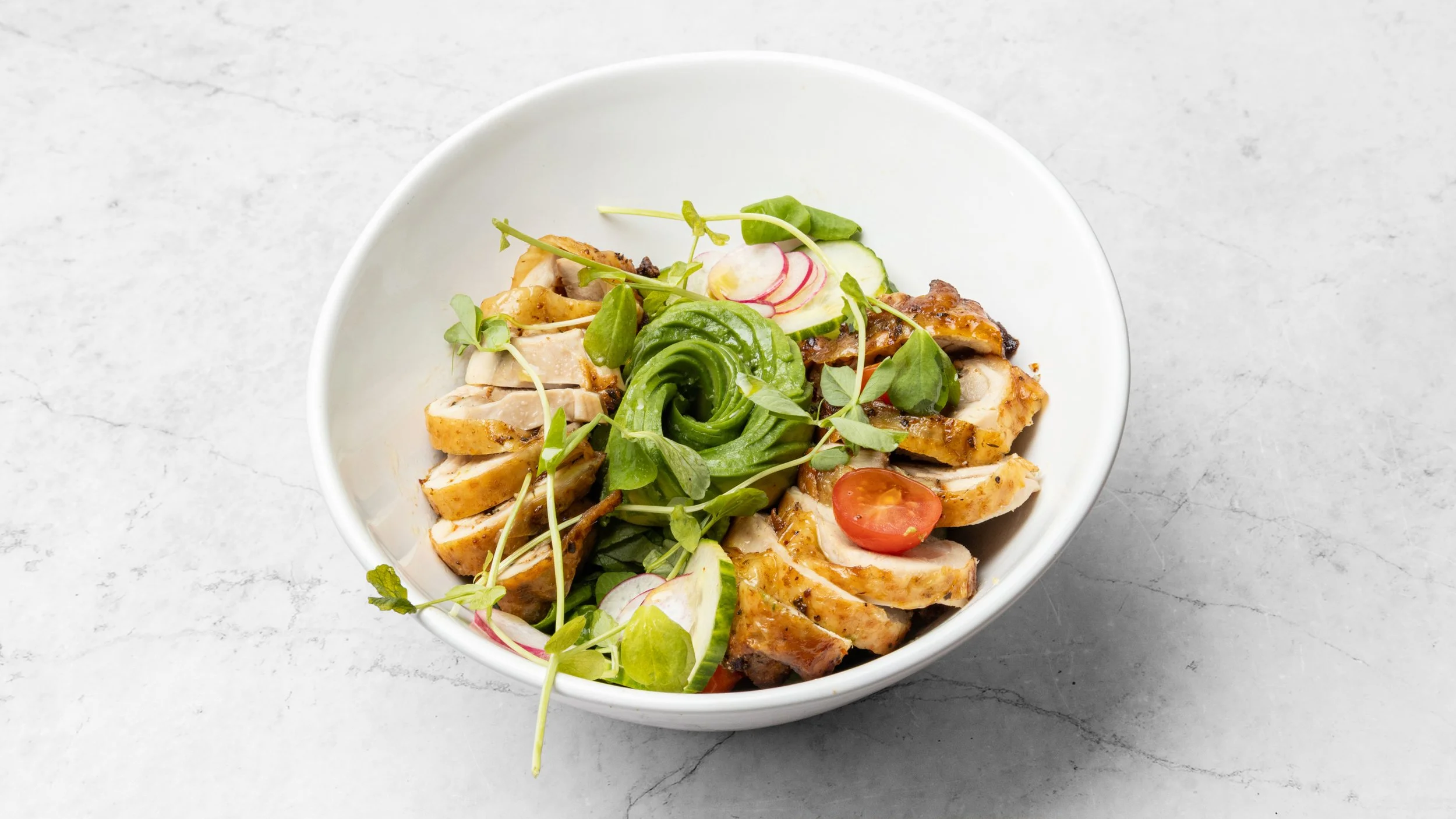 A white bowl with sliced grilled chicken, cherry tomatoes, radishes, cucumber slices, avocado slices, and microgreens on a light gray surface.