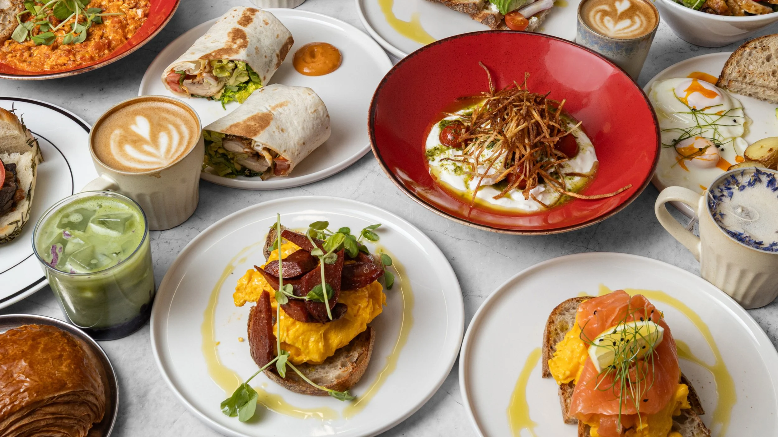A variety of Spanish-style dishes and beverages on a table, including tapas with smoked salmon, scrambled eggs with chorizo, hummus with vegetables, a green smoothie, coffee drinks with foam art, and a burrito.