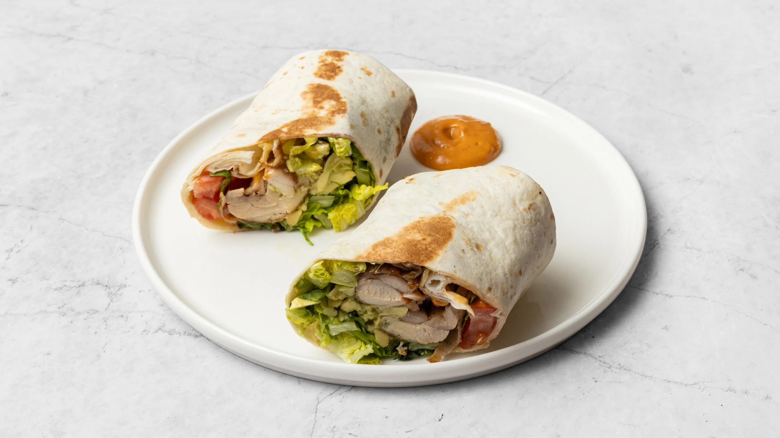 Two chicken wrap sandwiches on a white plate with a side of orange sauce on a light-colored surface.