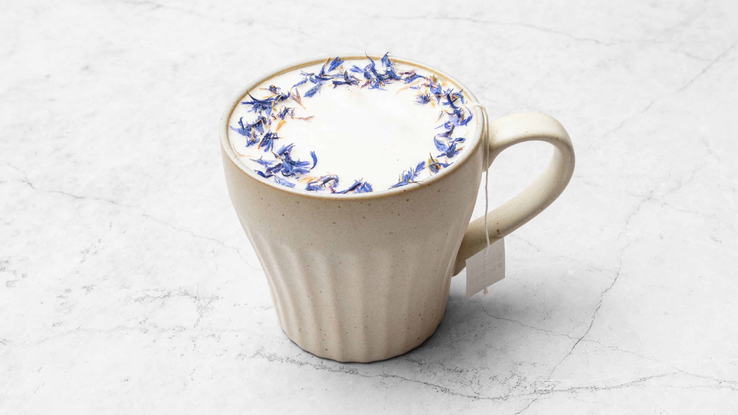 A beige ceramic mug filled with a frothy beverage topped with dried blue flower petals on a white marble surface.