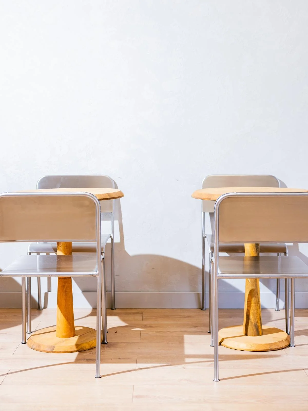Two beige chairs with metal frames and two light wooden tables in a minimalist setting against a plain white wall.