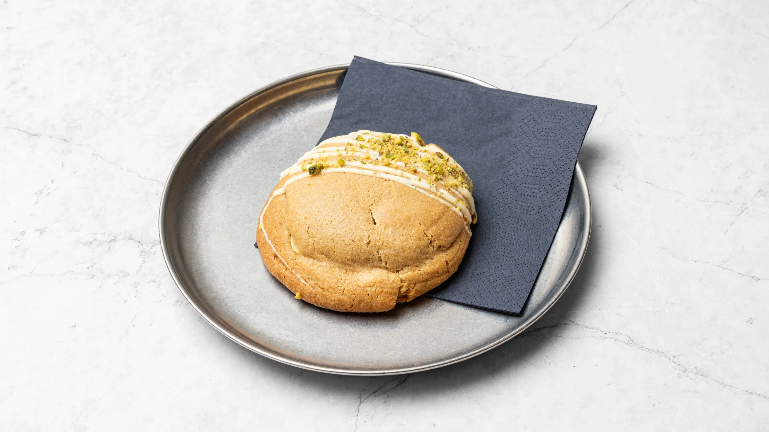A cookie with white icing and chopped pistachios on top, placed on a dark napkin on a silver plate, against a white marble surface.