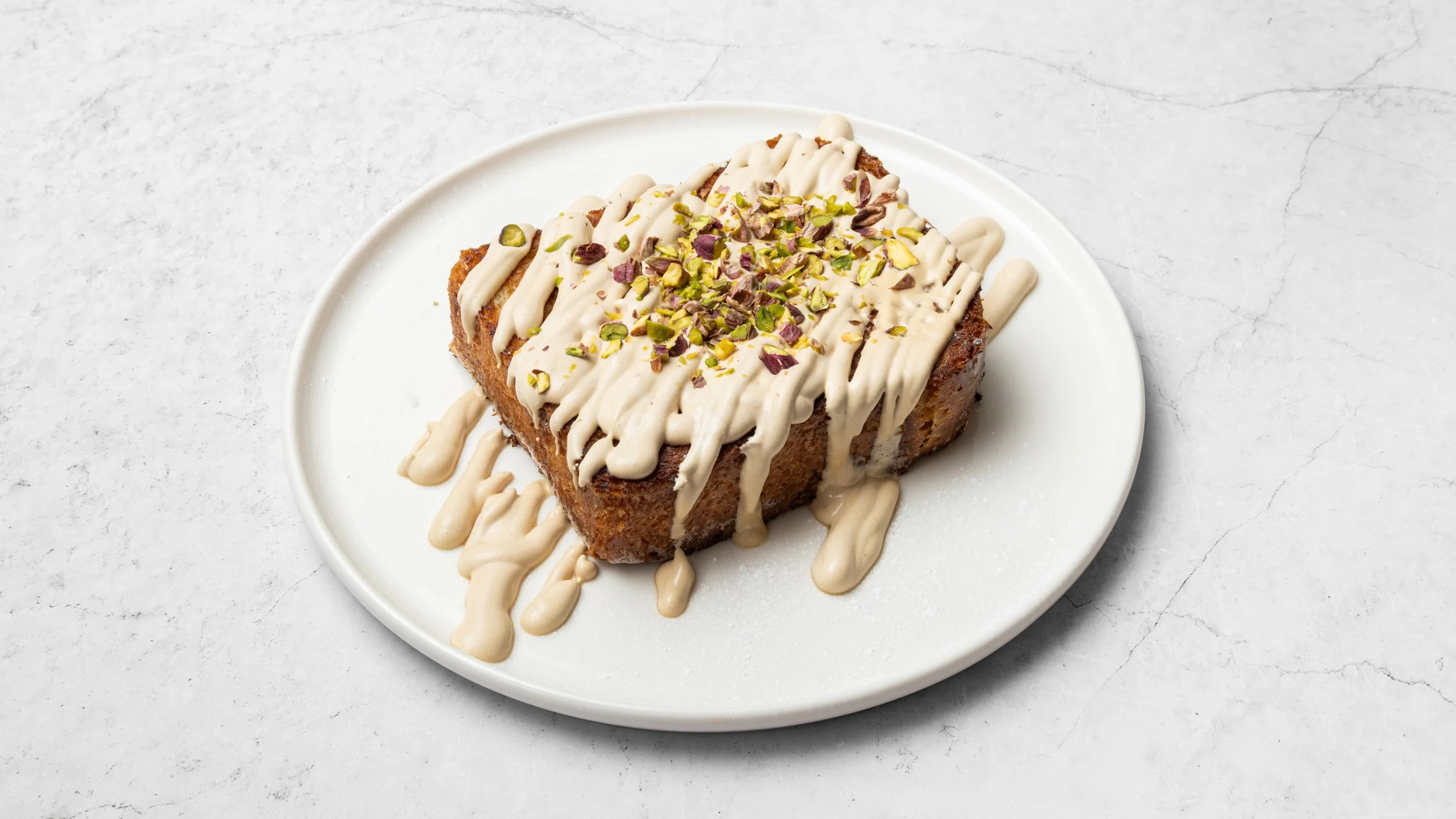 A slice of cake on a white plate, topped with white icing, chopped pistachios, and drizzled with icing.