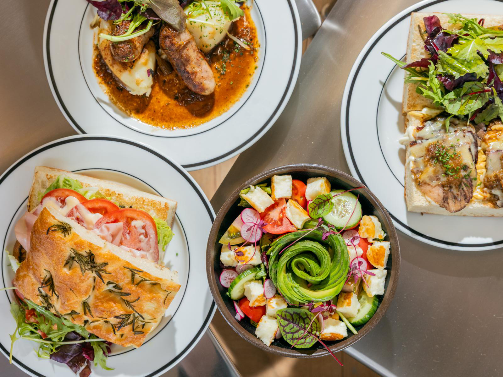 A top-down view of four plates of food and a salad bowl on a table. The plates include a meat dish with mashed potatoes and greens, a sandwich with tomato and ham, a sandwich with roasted meat and greens, and a spinach salad with cherry tomatoes, cucumbers, radishes, feta cheese, and microgreens.