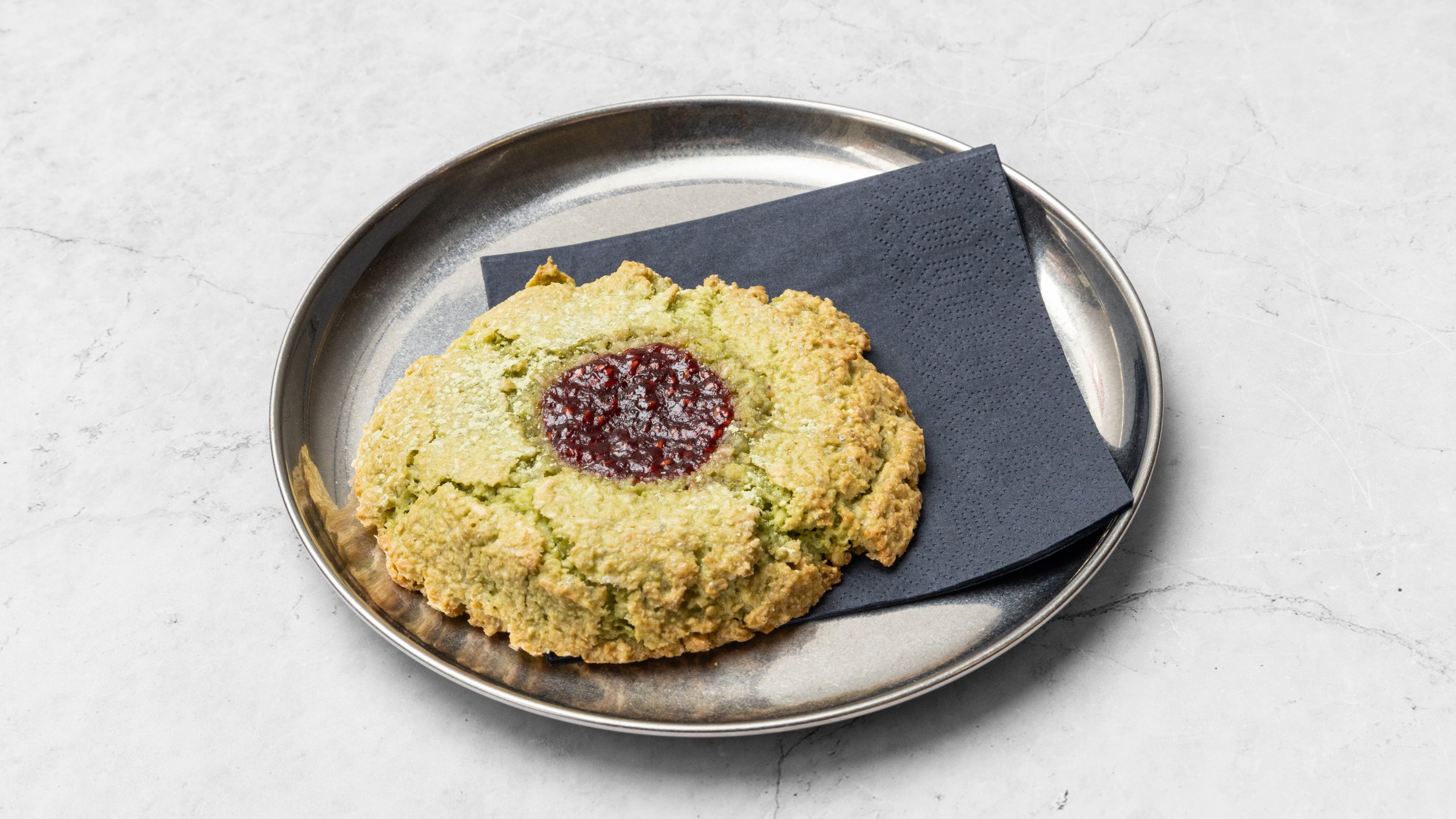 A slice of greenish crumble cake with a dollop of red jam on top, on a metal tray with a black napkin, on a white marbled surface.