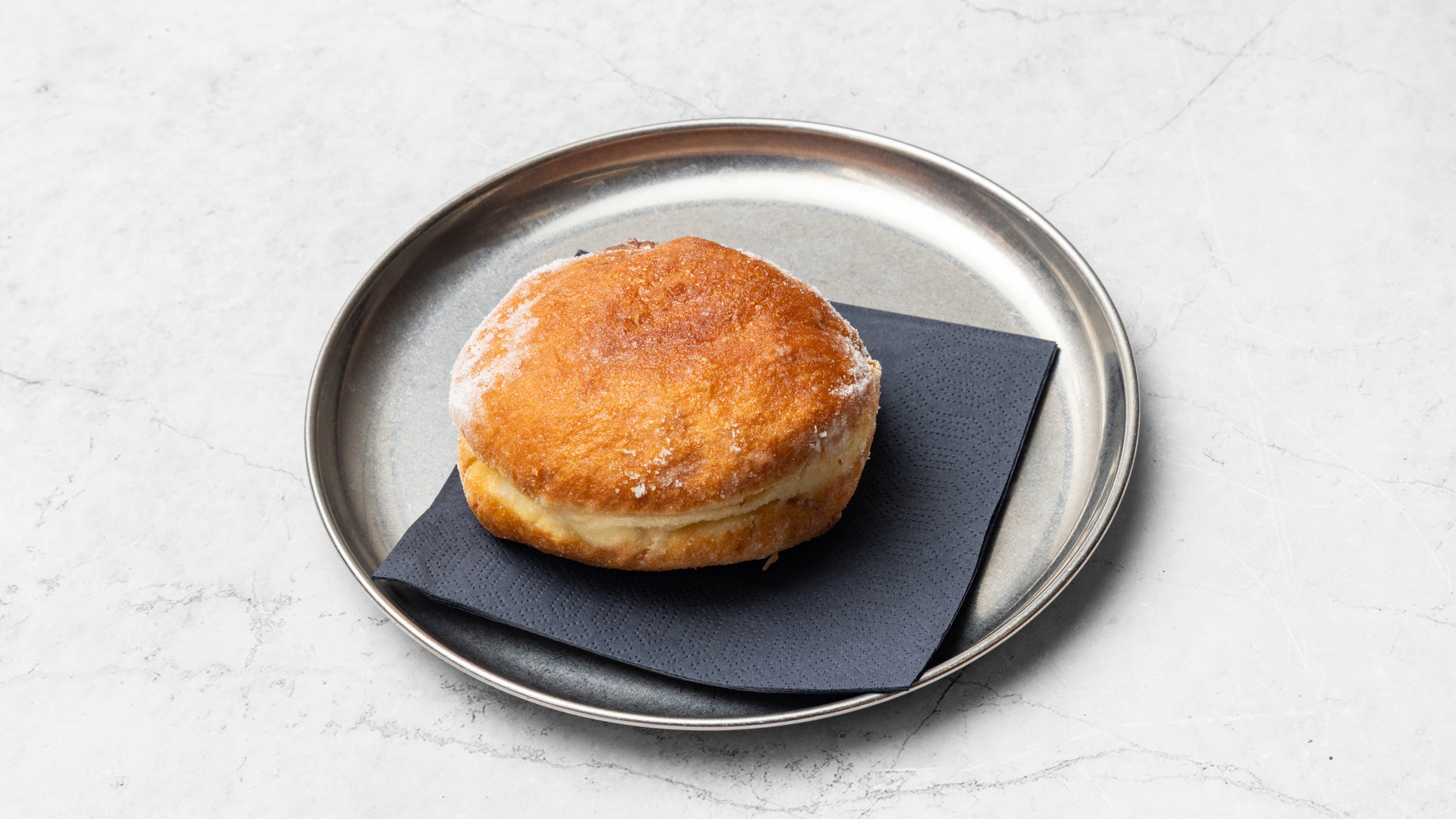 Fried biscuit with powdered sugar on a black napkin, placed on a round metal tray on a white marble surface.