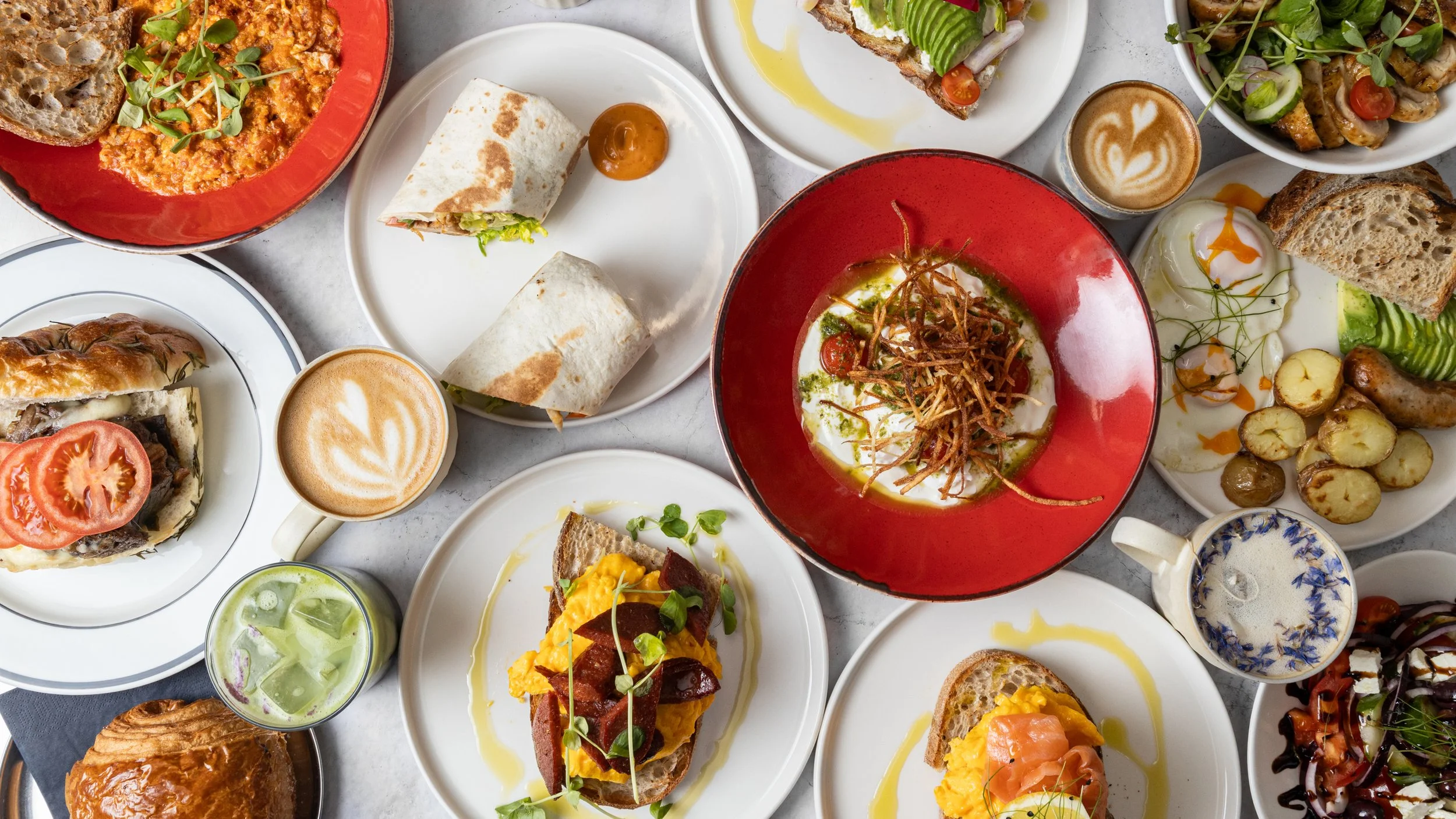 A spread of various dishes including toast, wraps, salads, poached eggs, coffee drinks, and side dishes on plates and cups.