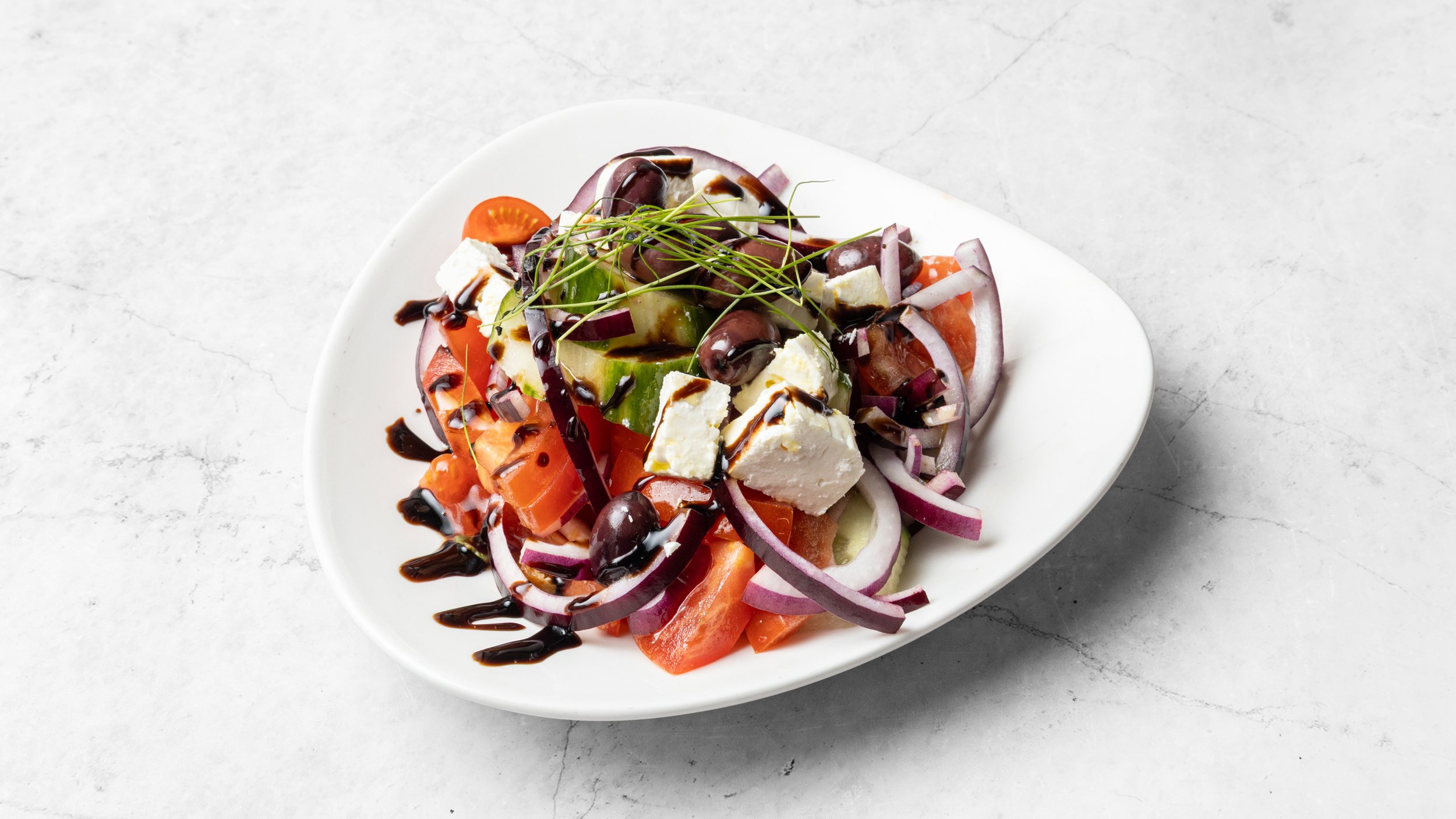 Greek salad with tomatoes, cucumbers, red onions, olives, feta cheese, and a balsamic glaze on a white oval plate.