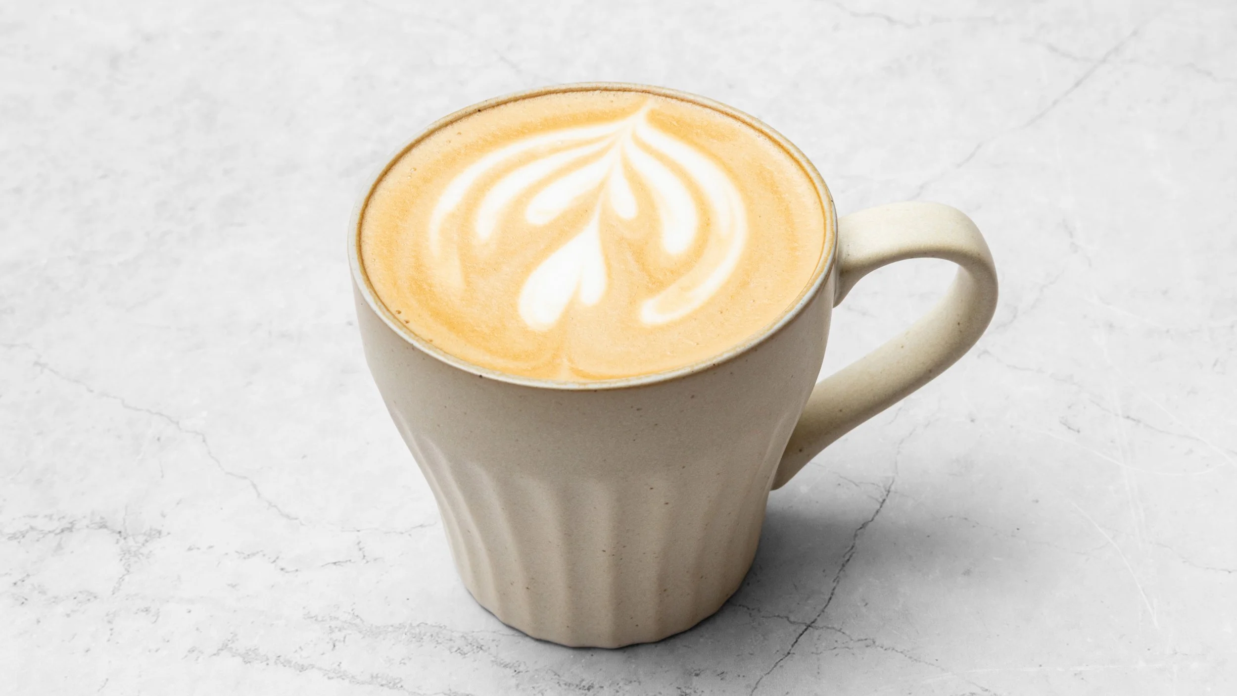 A beige ceramic mug filled with a latte displaying latte art on top, placed on a white marble surface.