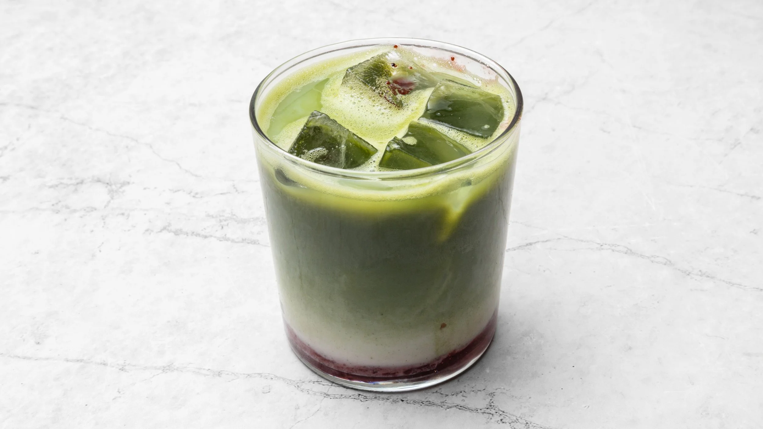 A tall glass of layered drink with green and red liquids, ice cubes, and foamy top, on a white marble surface