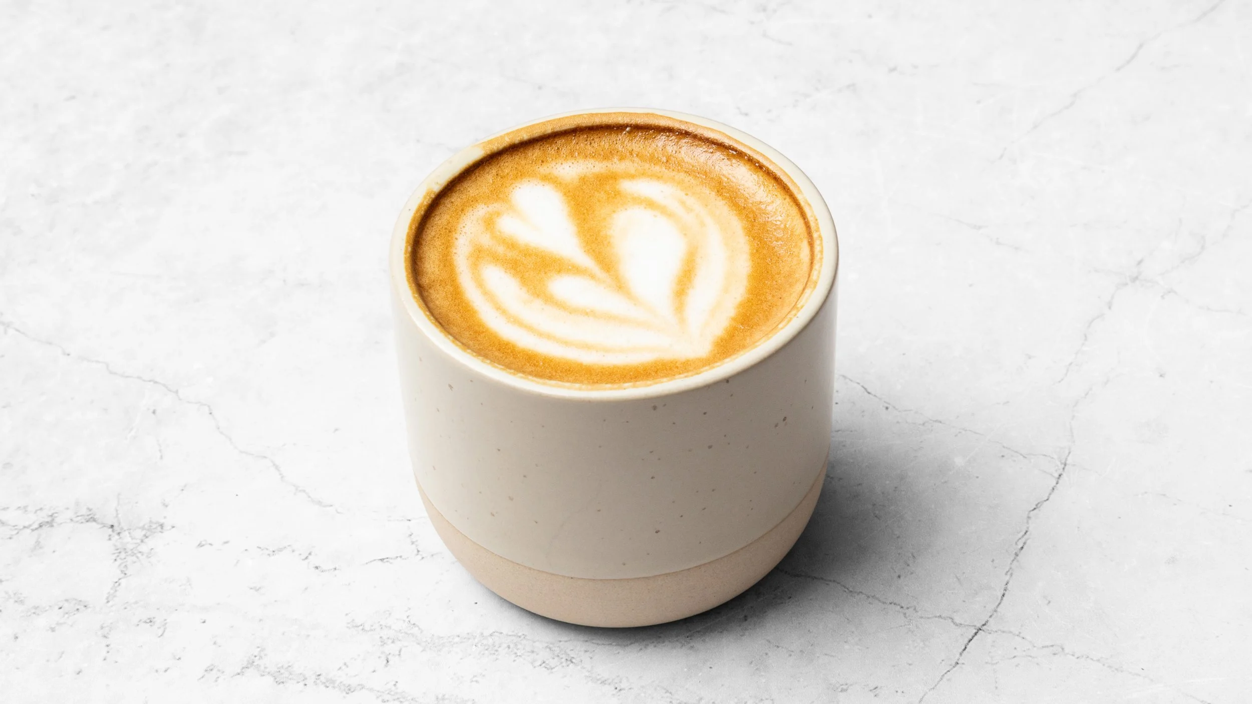 A white ceramic mug filled with a latte featuring a heart-shaped foam art design, placed on a light-colored marble surface.