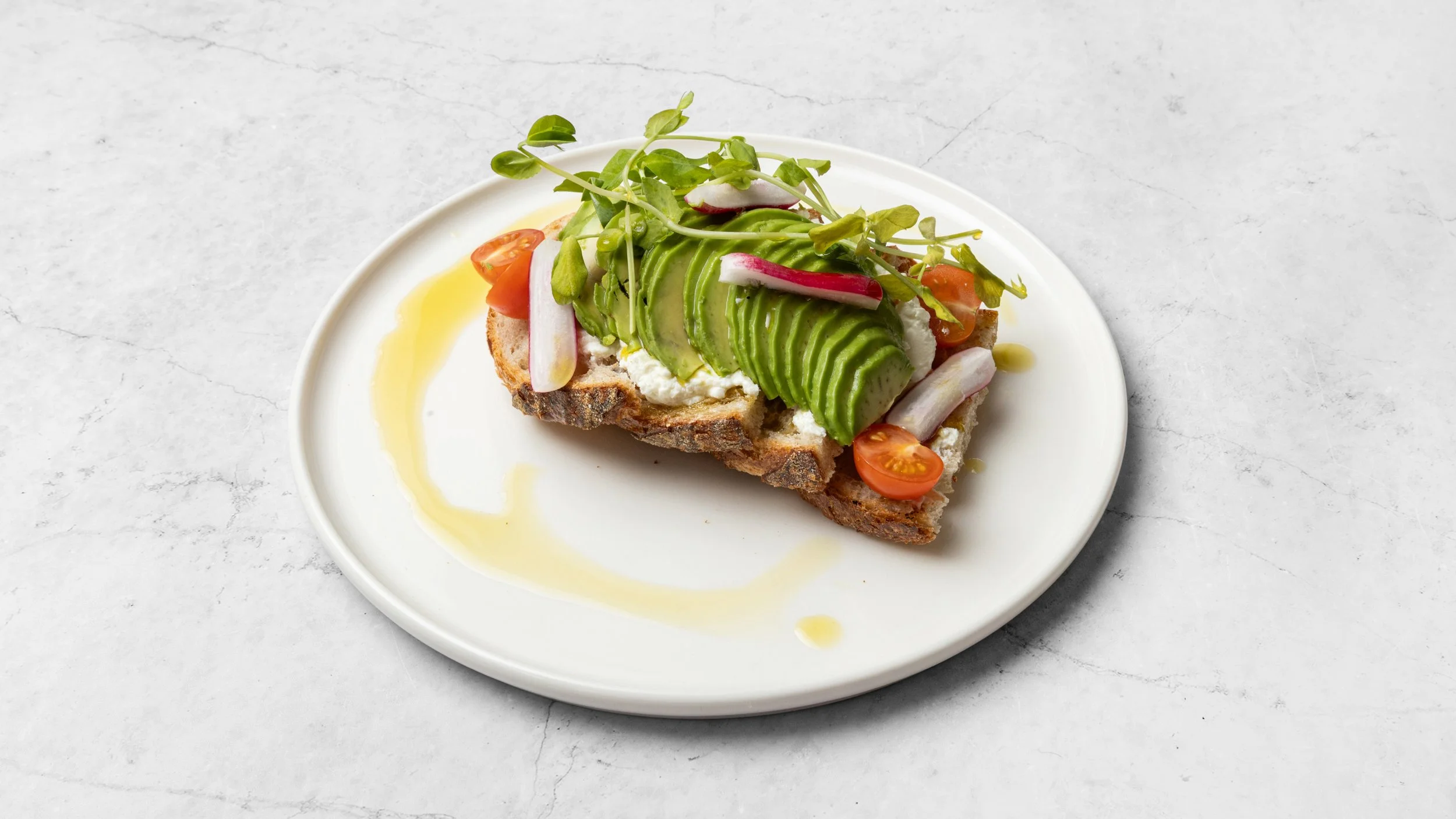 Open-faced sandwich on a slice of toasted bread topped with sliced avocado, cherry tomato halves, radish slices, microgreens, and a drizzle of olive oil.