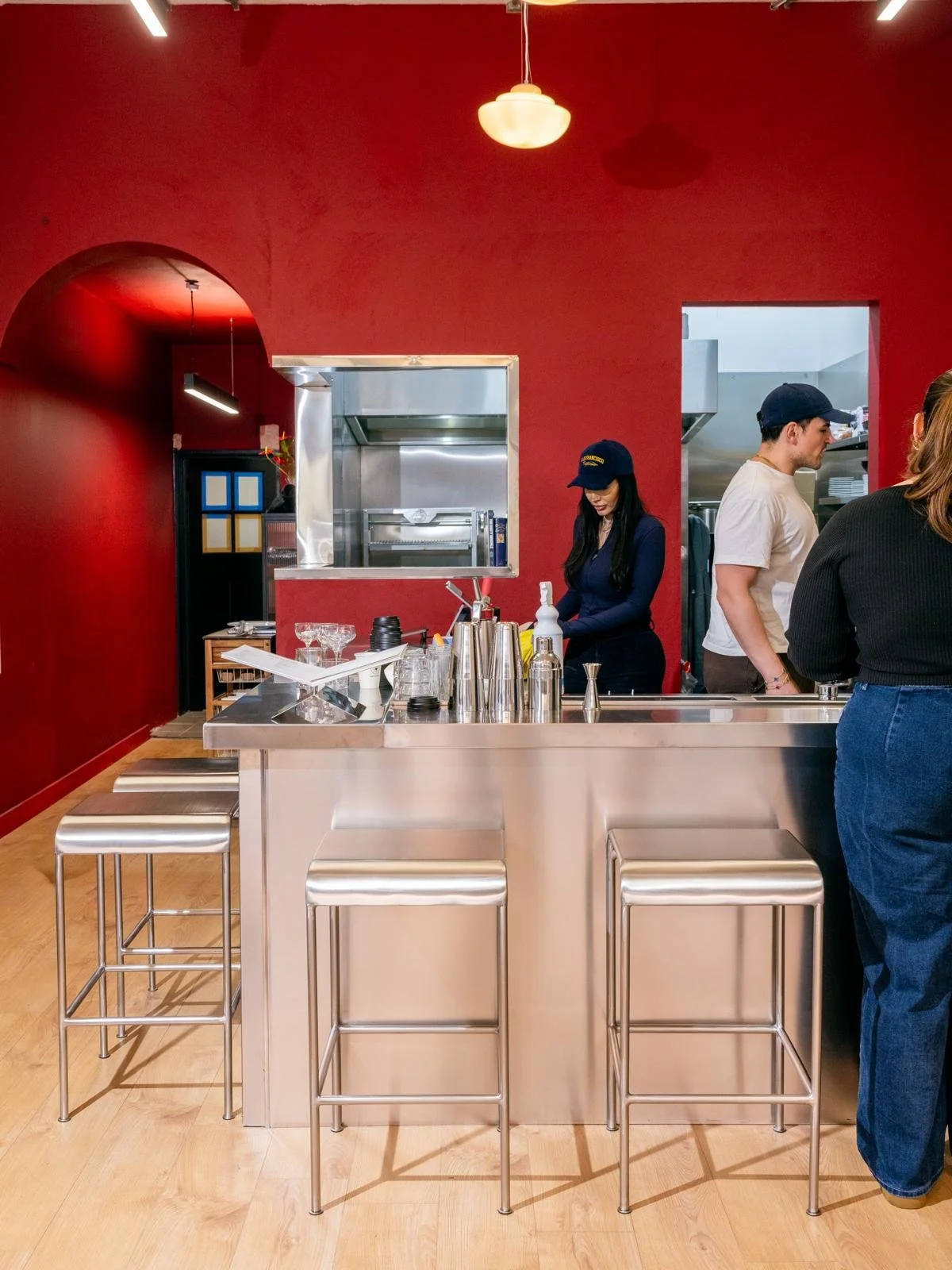 Part of a café or restaurant interior with a stainless steel counter, bar stools, and three people working behind the counter in front of a red wall.