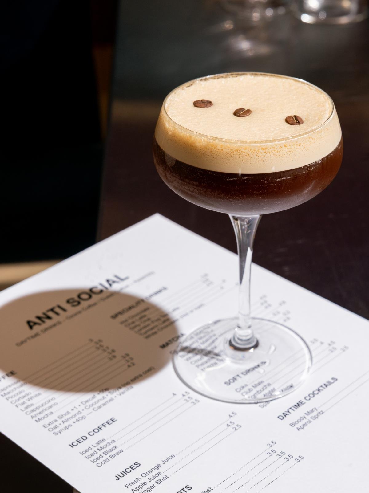 A cocktail glass filled with a dark beverage topped with a creamy foam and coffee beans, placed on an anti-social club drink menu.