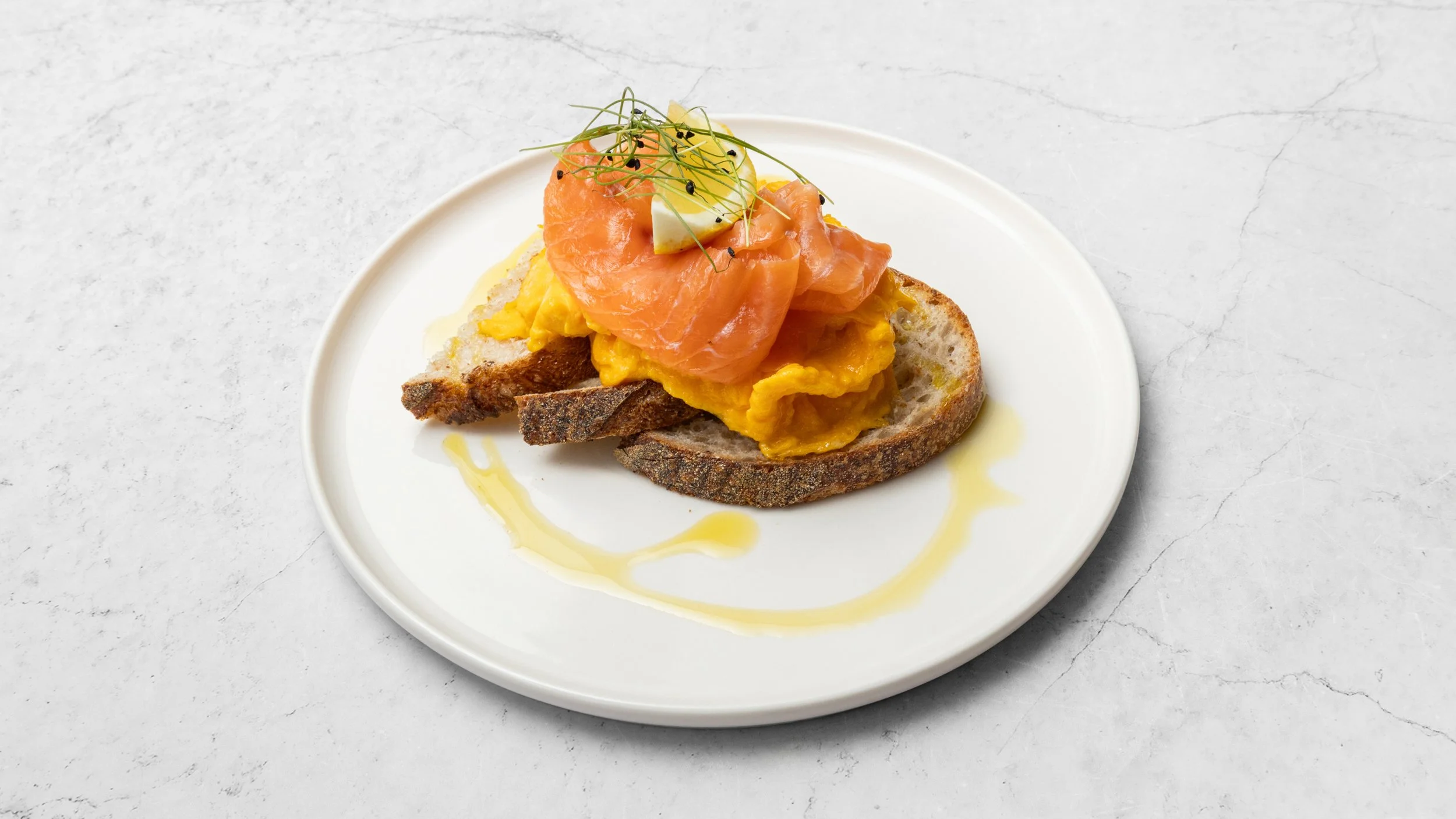 Slice of toasted bread topped with scrambled eggs, smoked salmon, a boiled egg quarter, and garnished with microgreens on a white plate with a drizzle of yellow sauce.