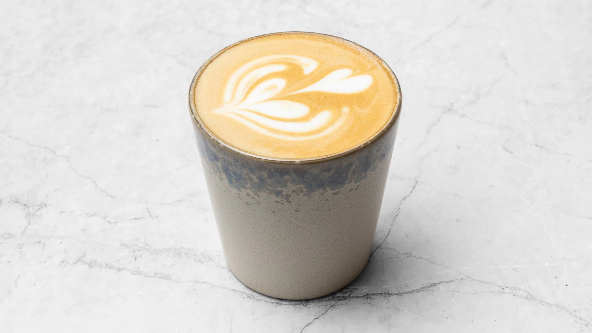 A tall glass of coffee with latte art on top, placed on a white marble surface.