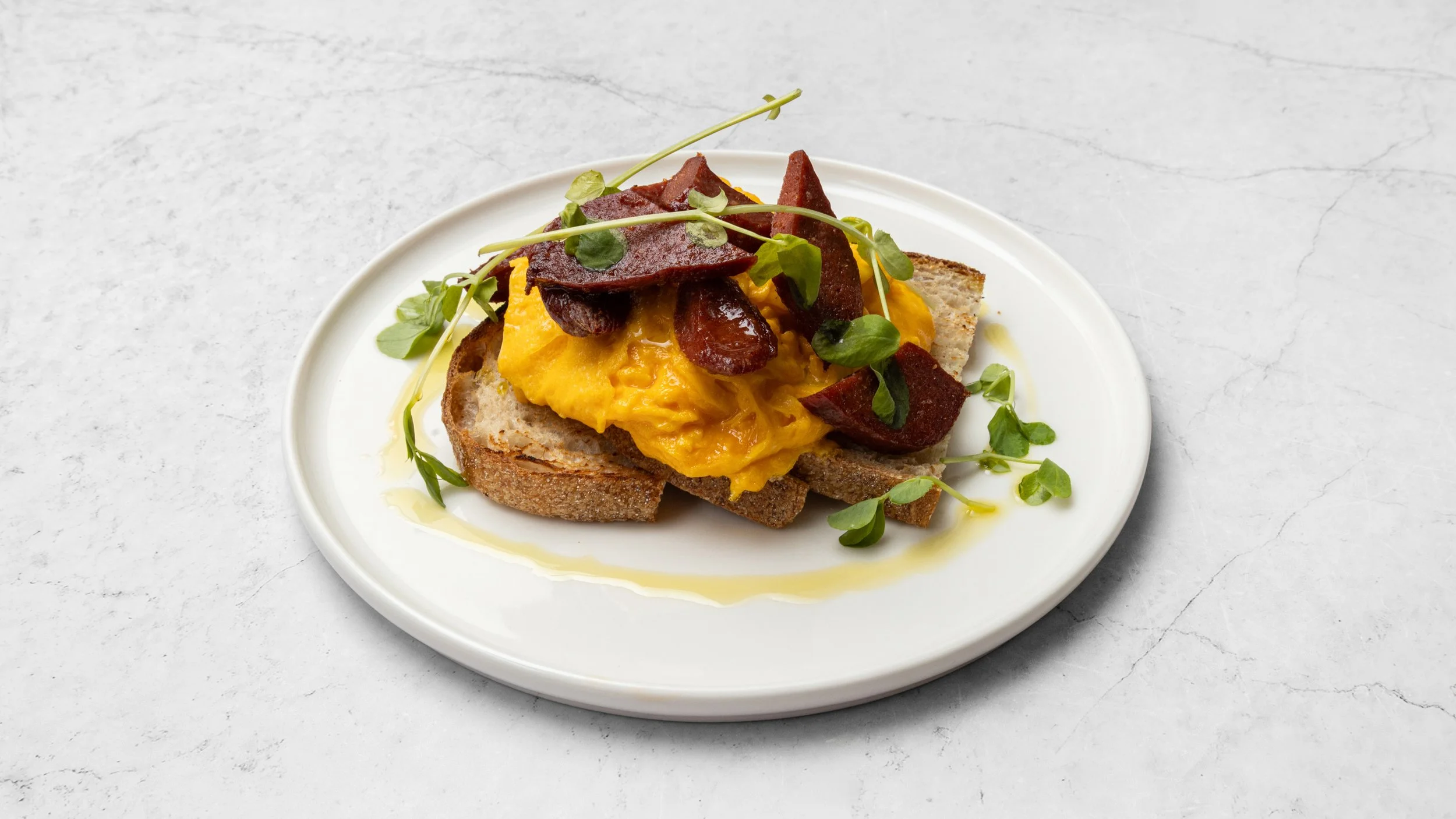 Open-faced sandwich with scrambled eggs, crispy bacon, microgreens, and olive oil on toasted bread served on a white plate.
