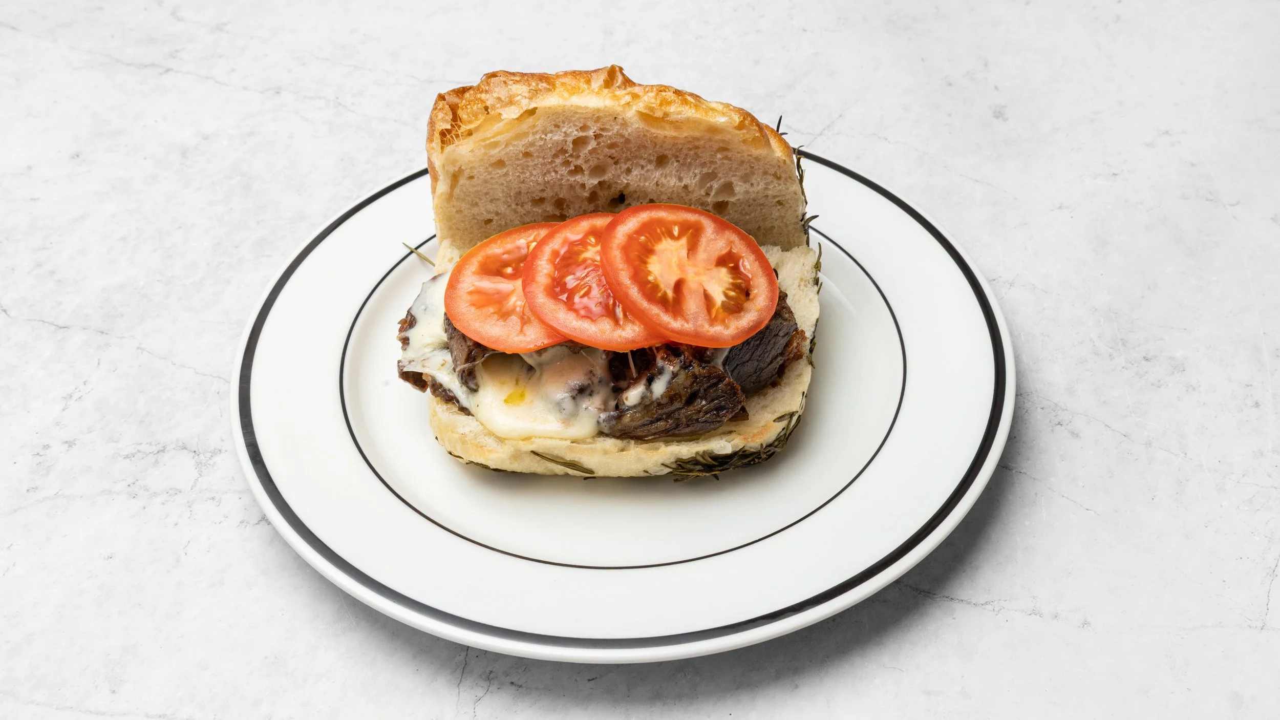 Open sandwich with melted cheese, cooked beef, and tomato slices on a white plate with black trim, placed on a light gray surface.