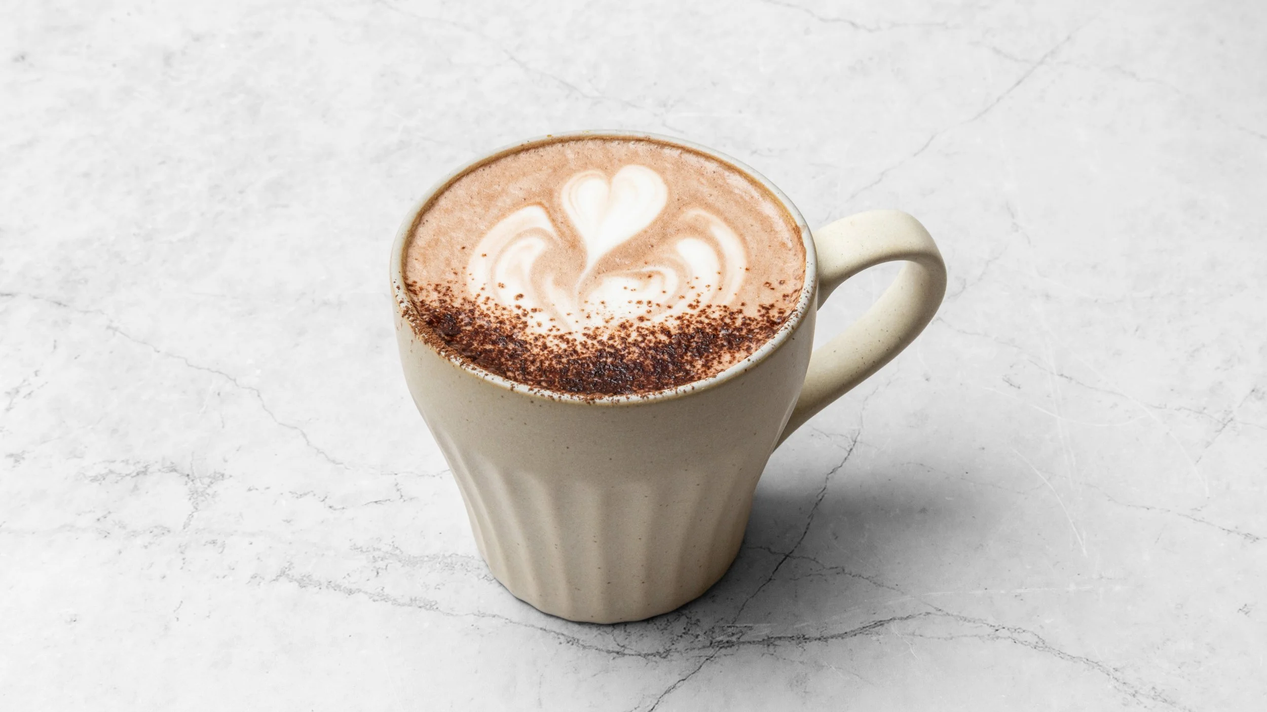 A ceramic mug with a frothy coffee drink decorated with latte art in the shape of a heart and surrounded by a sprinkle of cocoa powder.