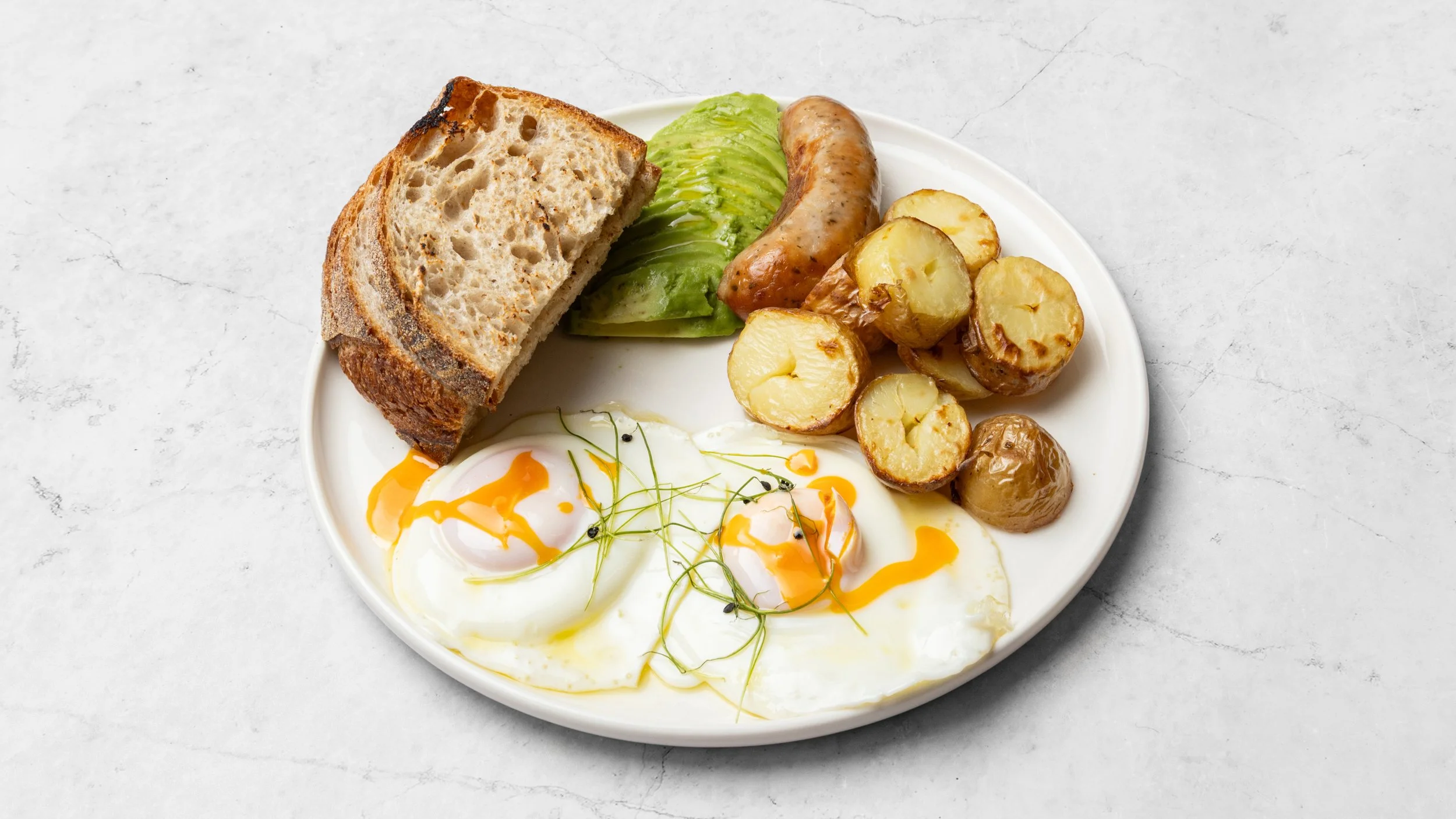 Plate of breakfast food including toasted bread, avocado slices, sausage, roasted potatoes, and eggs with yolk drizzled with sauce, garnished with herbs.