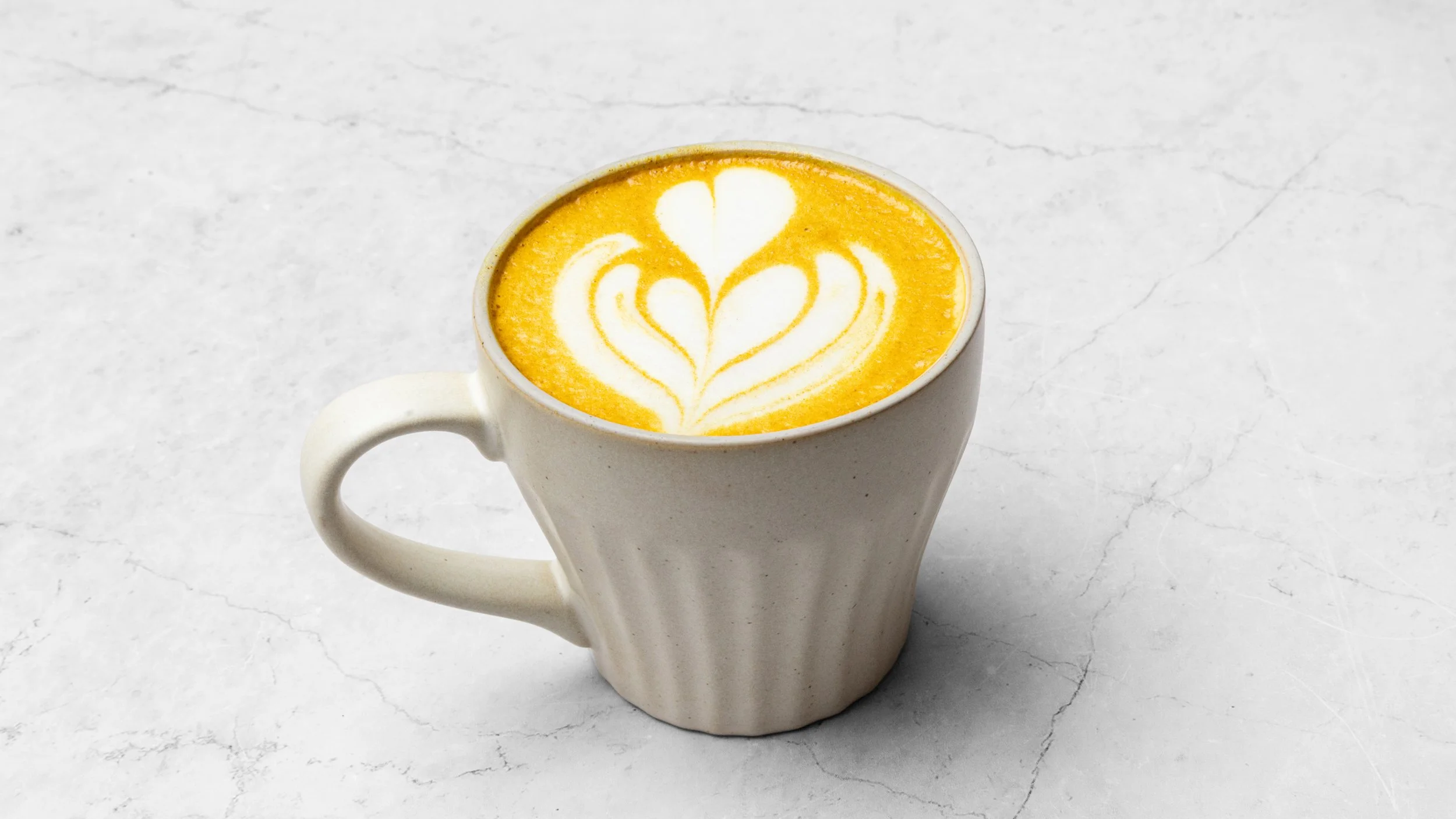 A white ceramic mug filled with a yellow latte featuring foam art in the shape of a heart and floral designs, placed on a light gray marble surface.