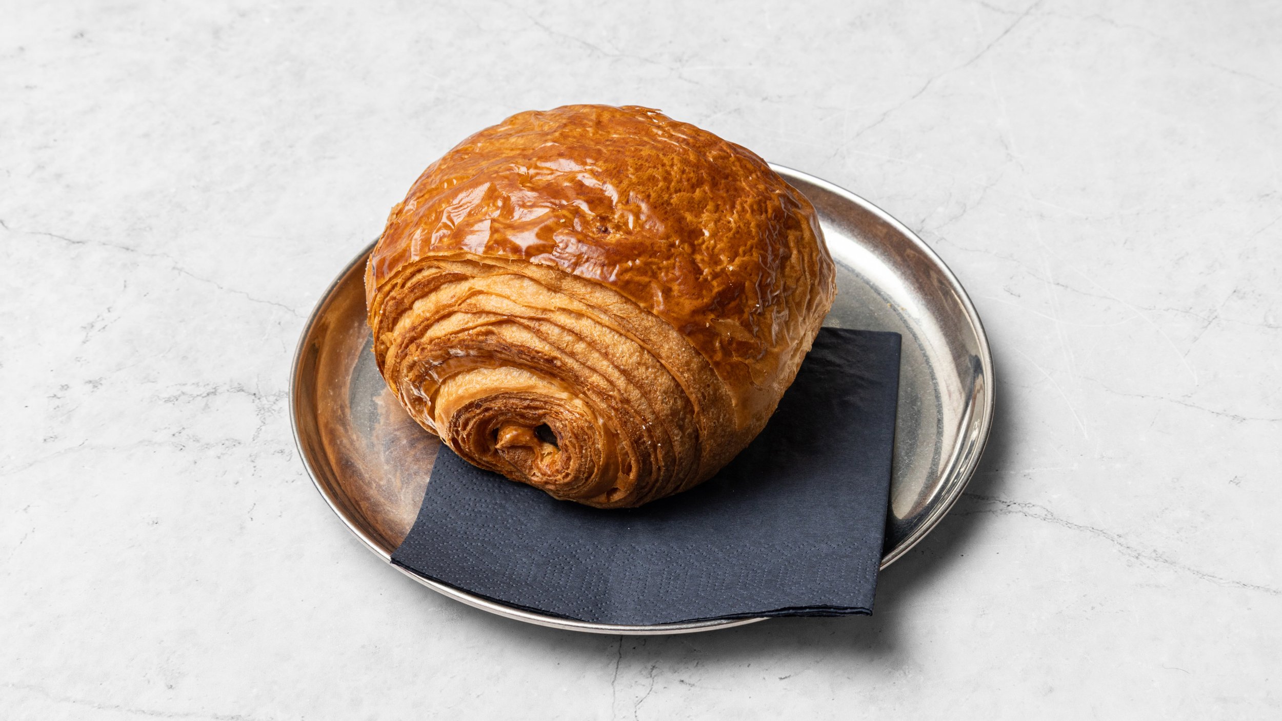 A large, golden-brown croissant with a shiny glaze sits on a silver tray lined with a black napkin, on a light gray marble surface.
