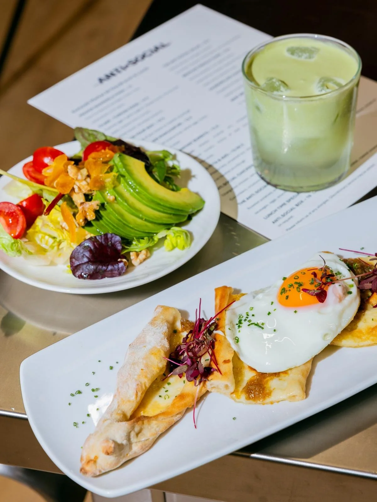 Salad with tomatoes, avocado, lettuce, and nuts, a glass of iced green drink, and a plate of flatbread topped with a fried egg and garnishes on a table with a menu in the background.