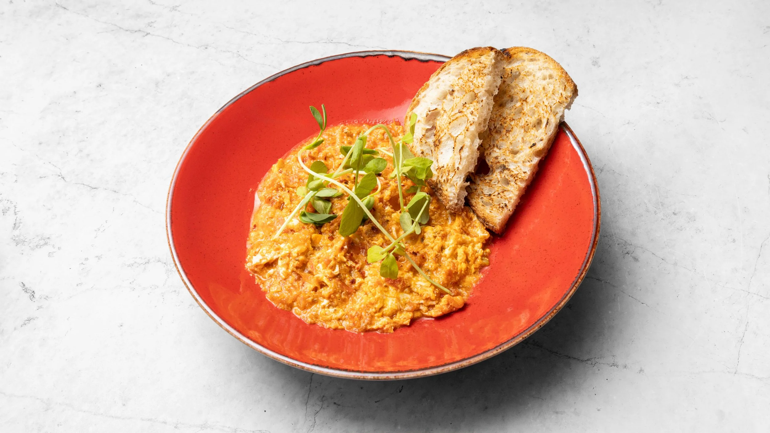 Bowl of scrambled eggs garnished with microgreens, served with slices of toasted bread on a red plate.