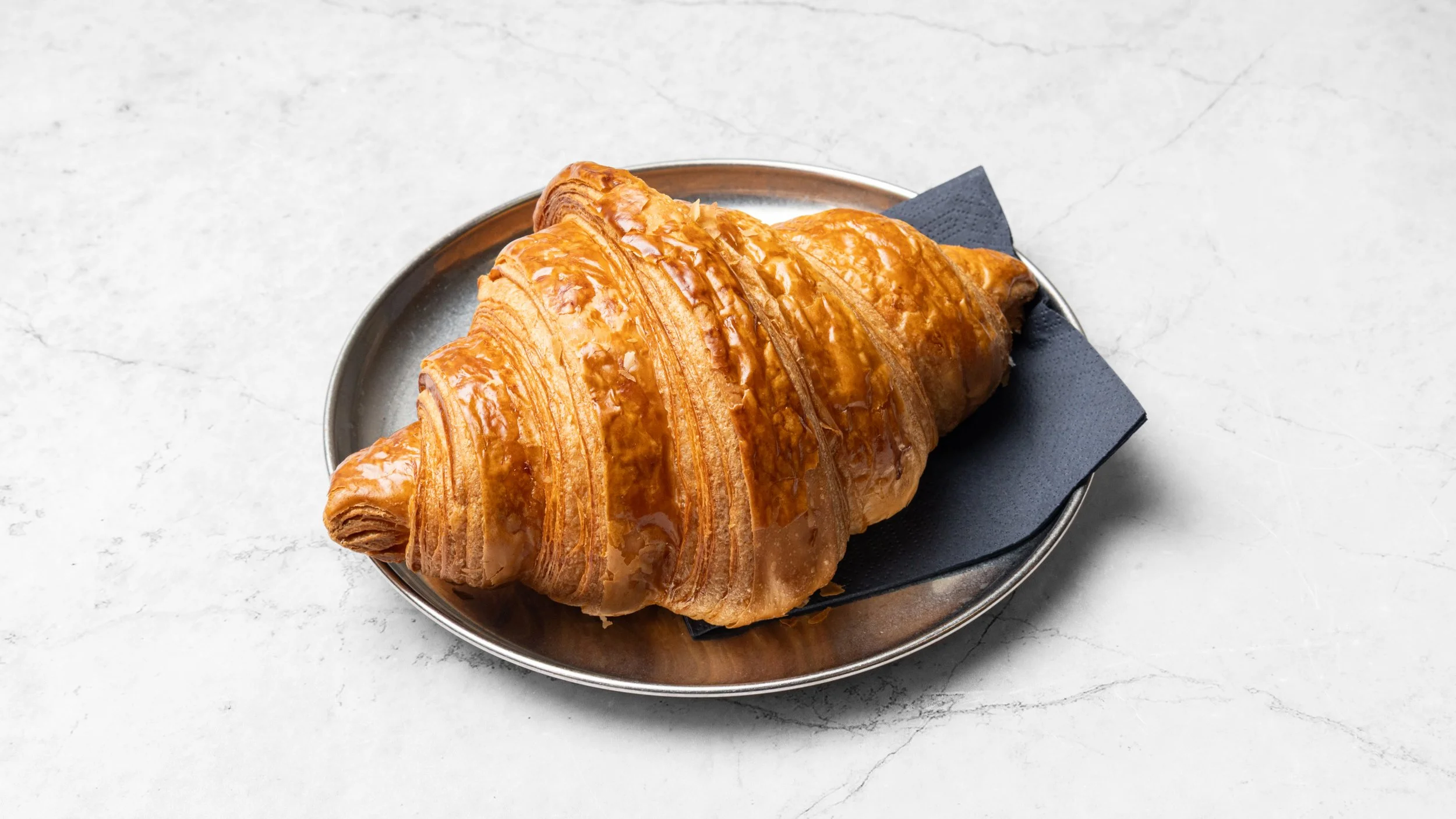 Golden-brown croissant with a glossy glaze on a metal plate, placed on a black napkin on a white marble surface.
