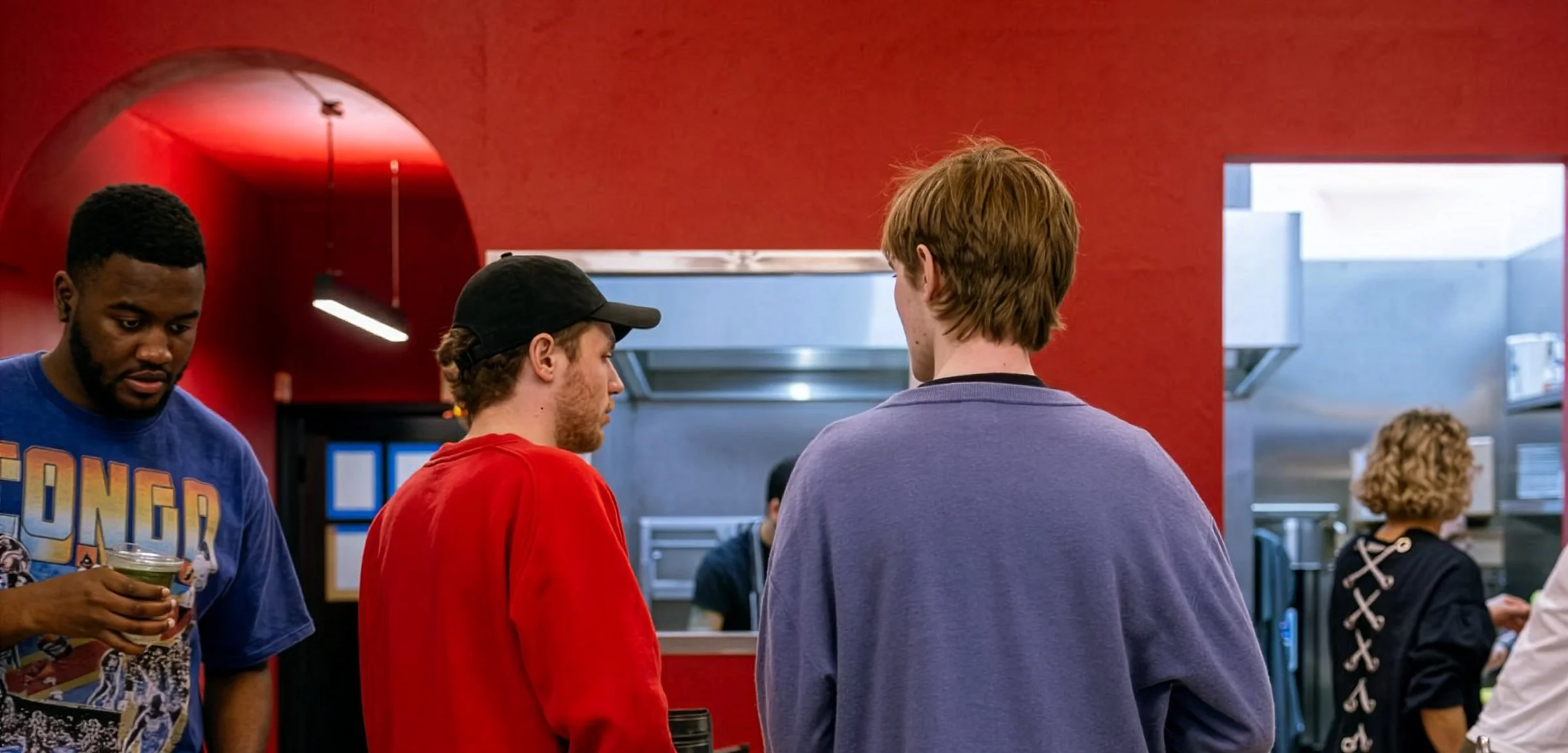 Three young men standing in a line inside a fast food restaurant. One with a blue T-shirt, one with a red hoodie, and one with a purple sweater, are engaged in conversation. There are other people in the background, and the restaurant has a red wall with an entrance to the kitchen area.