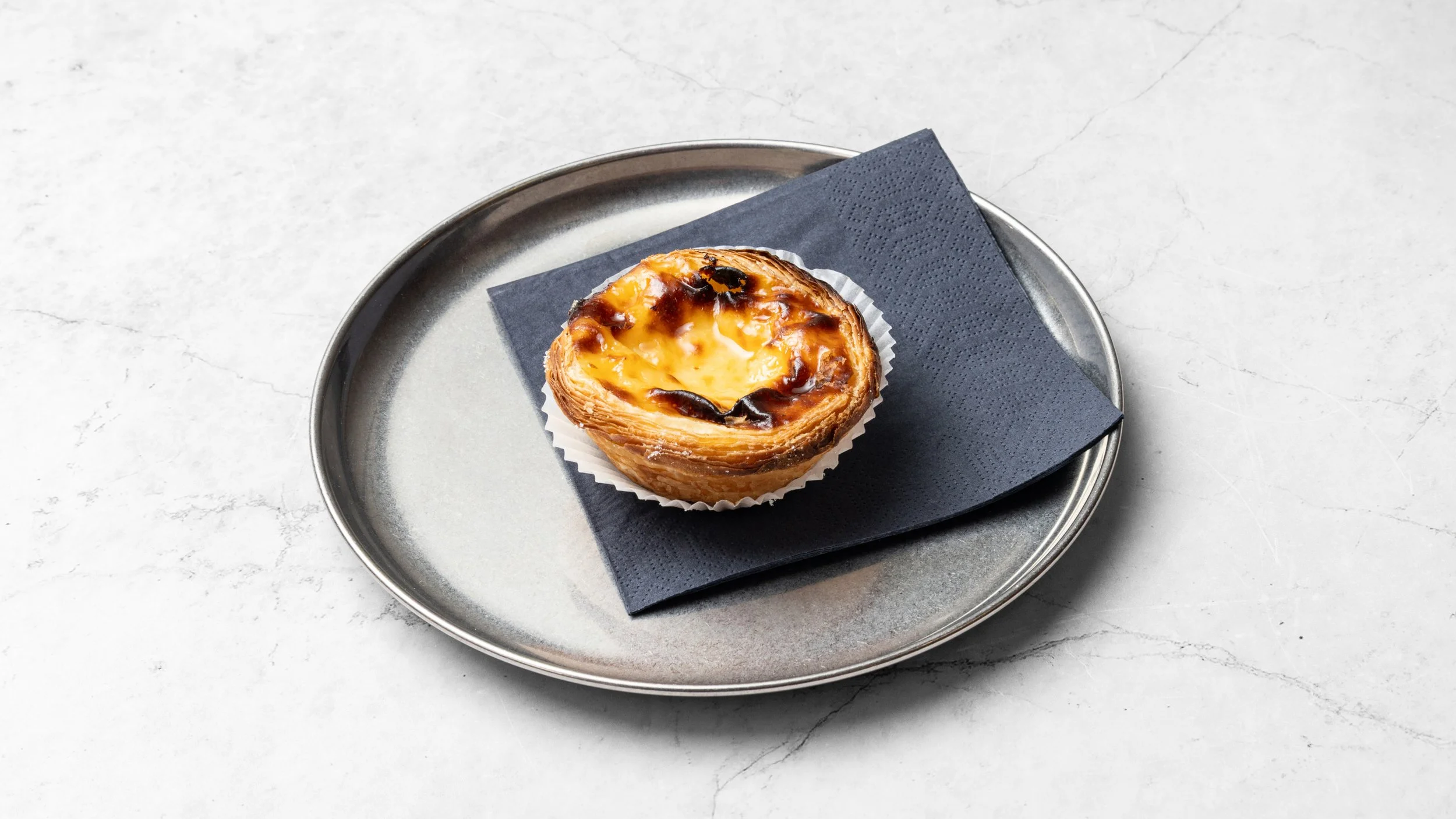 A Portuguese custard tart with a flaky crust and caramelized top on a metal tray, with a black napkin underneath, on a white marble surface.