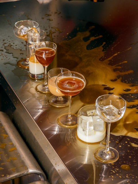 A row of cocktail glasses and glasses with ice, some with liquid and others with milk or cream, on a stainless steel surface with spilled liquid around them.