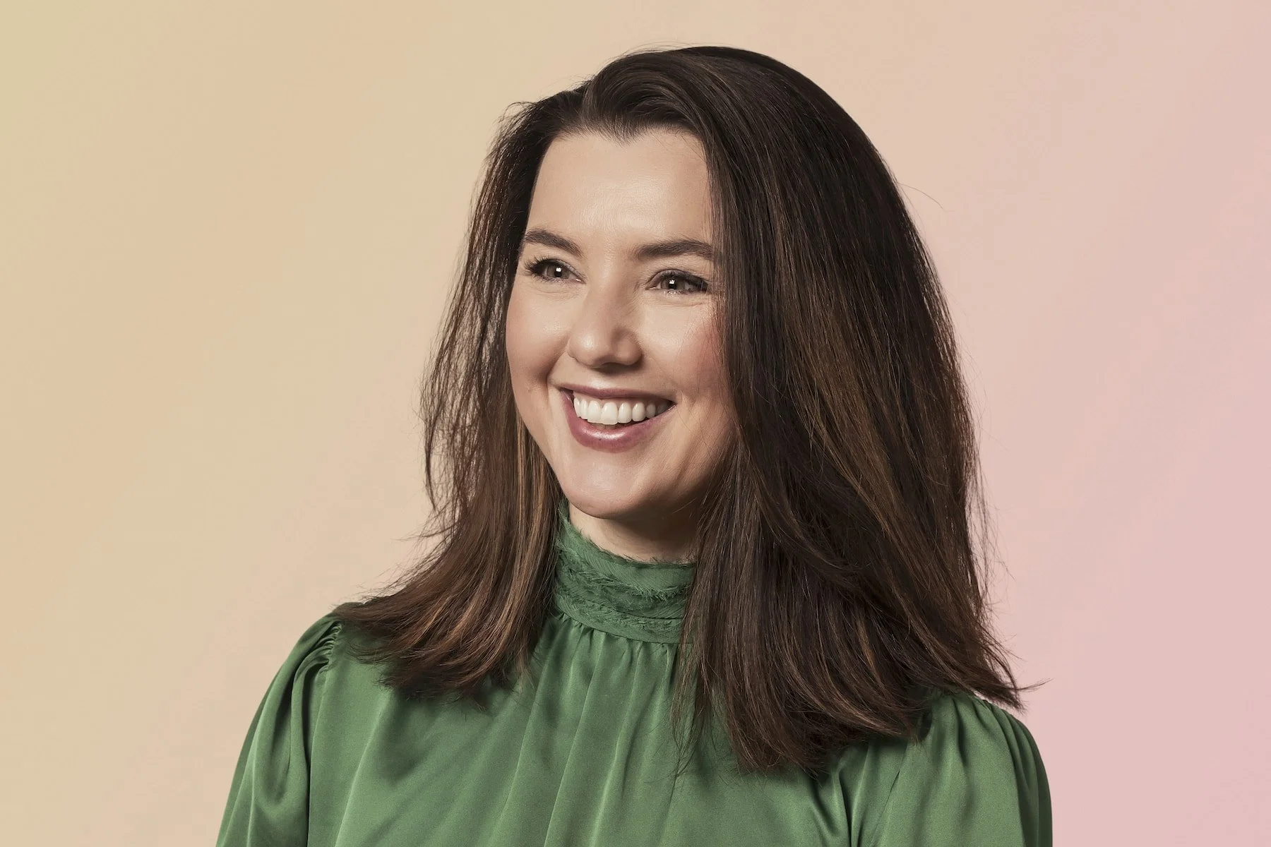 A smiling woman with long brown hair wearing a green top, standing against a pastel pink and beige background.