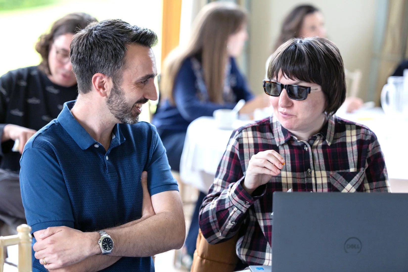 Photo of a woman wearing dark glasses talking to a man with a beard. They are smiling and collaborating on work with a laptop at an event.
