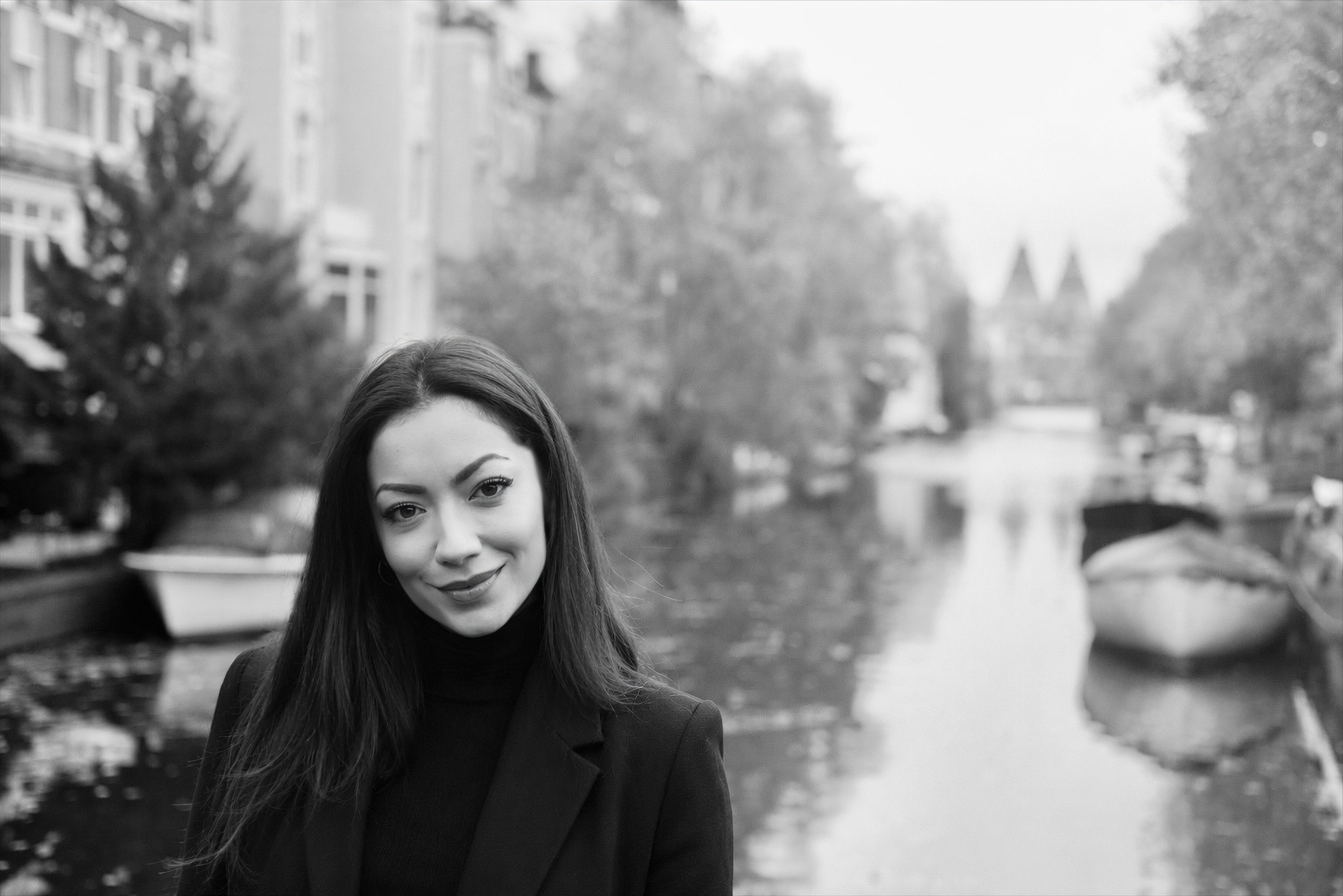 Porträt einer jungen Frau mit langen dunklen Haaren, schwarzer Kleidung und einem freundlichen Lächeln, im Freien an einem bewölkten Tag, mit Wasser und Bäumen im Hintergrund.