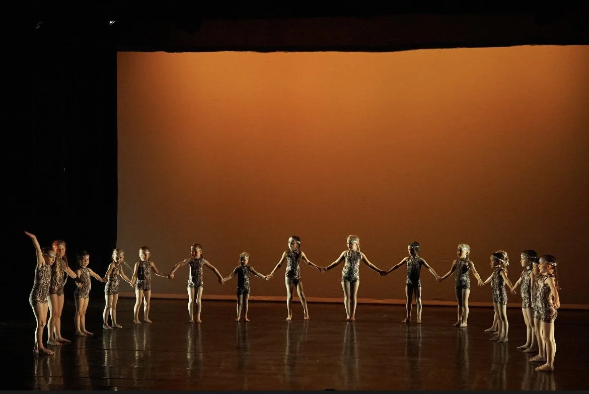 Children in dance costumes holding hands in a circle on stage during a performance.